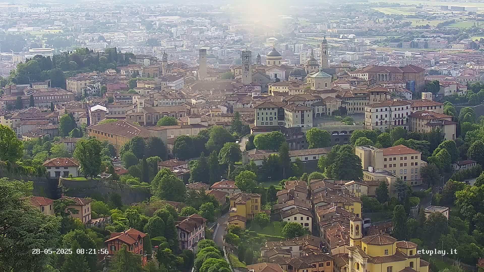 A hazy, high-angle view shows a sprawling town nestled amongst lush green trees and hills.