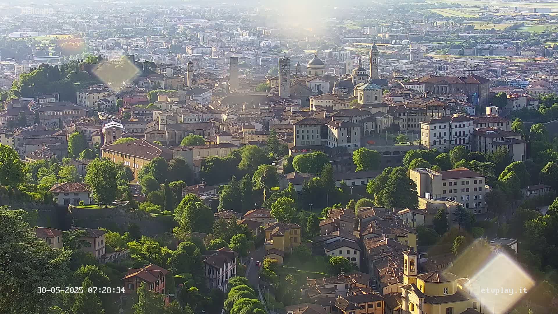 A sunlit city, nestled among lush green trees and hills, features numerous buildings of varying heights and architectural styles.