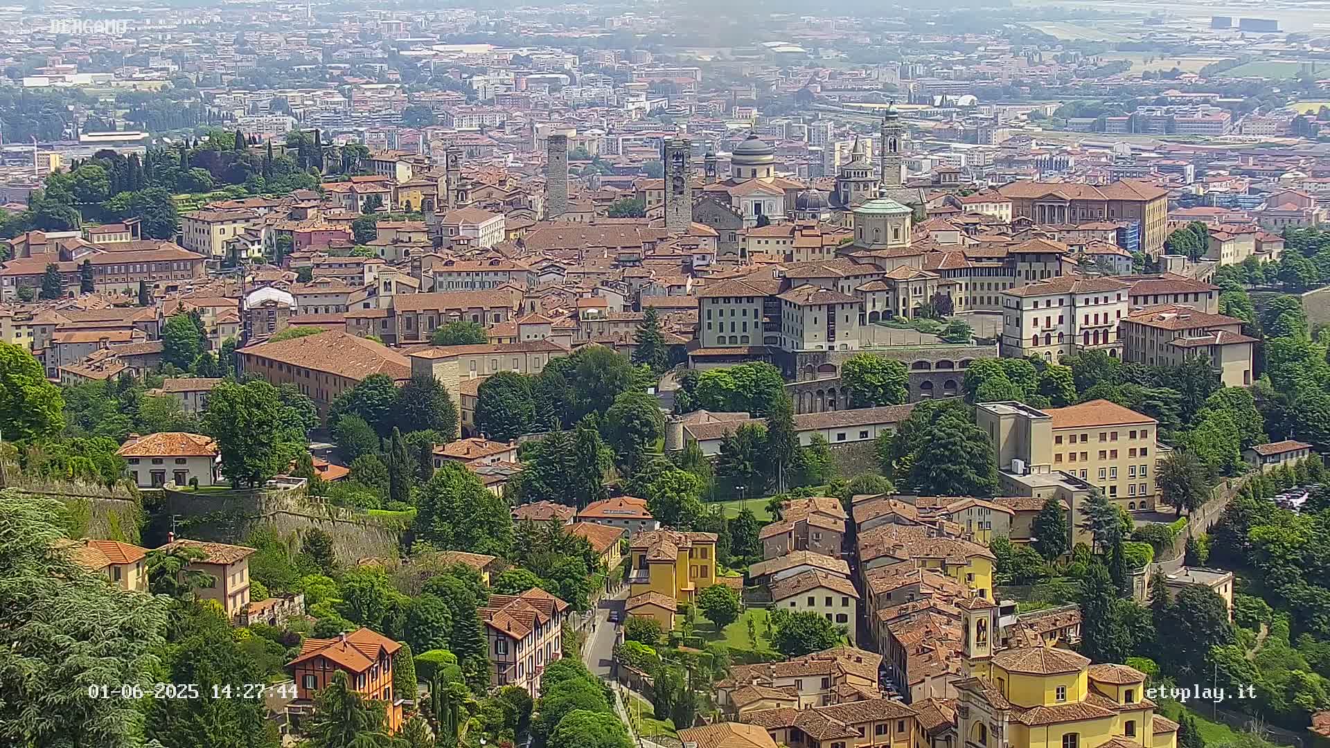 A sprawling, historic city with terracotta rooftops and numerous towers and churches is viewed from above on a hazy, sunny day.