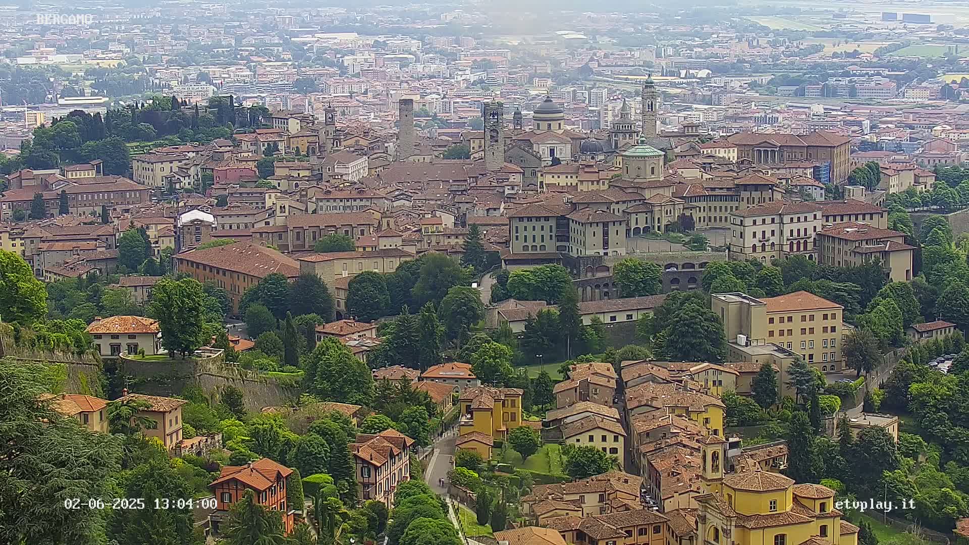 A sprawling city of terracotta-roofed buildings nestled among green trees and hills, under a hazy sky.