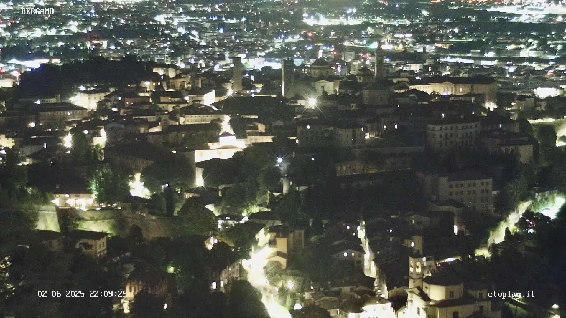 A nighttime aerial view shows a city with illuminated buildings and streets nestled within a landscape of dark green trees.