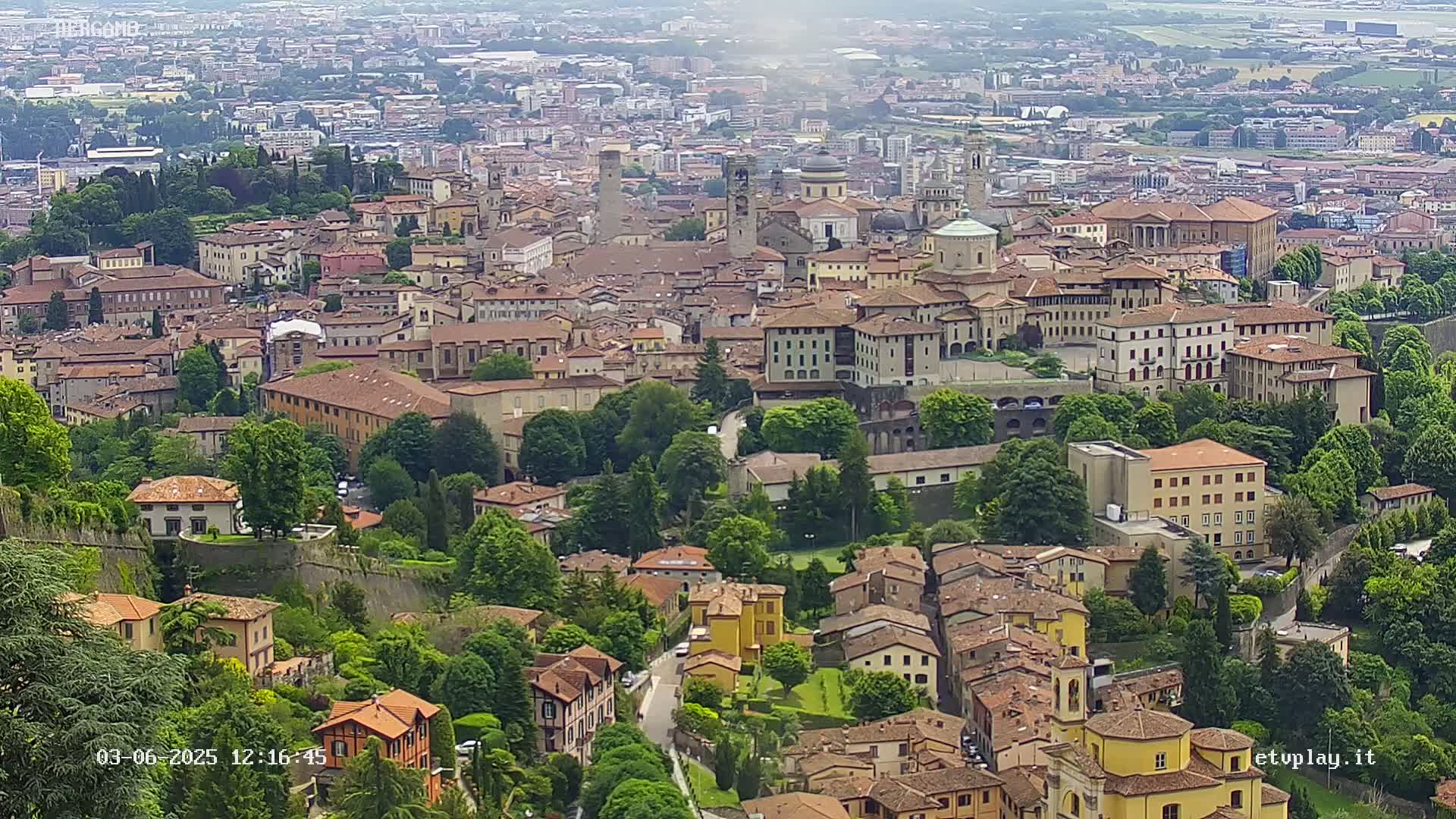 An aerial view of a dense, historic city with red-tiled roofs and lush green vegetation, under hazy weather conditions.