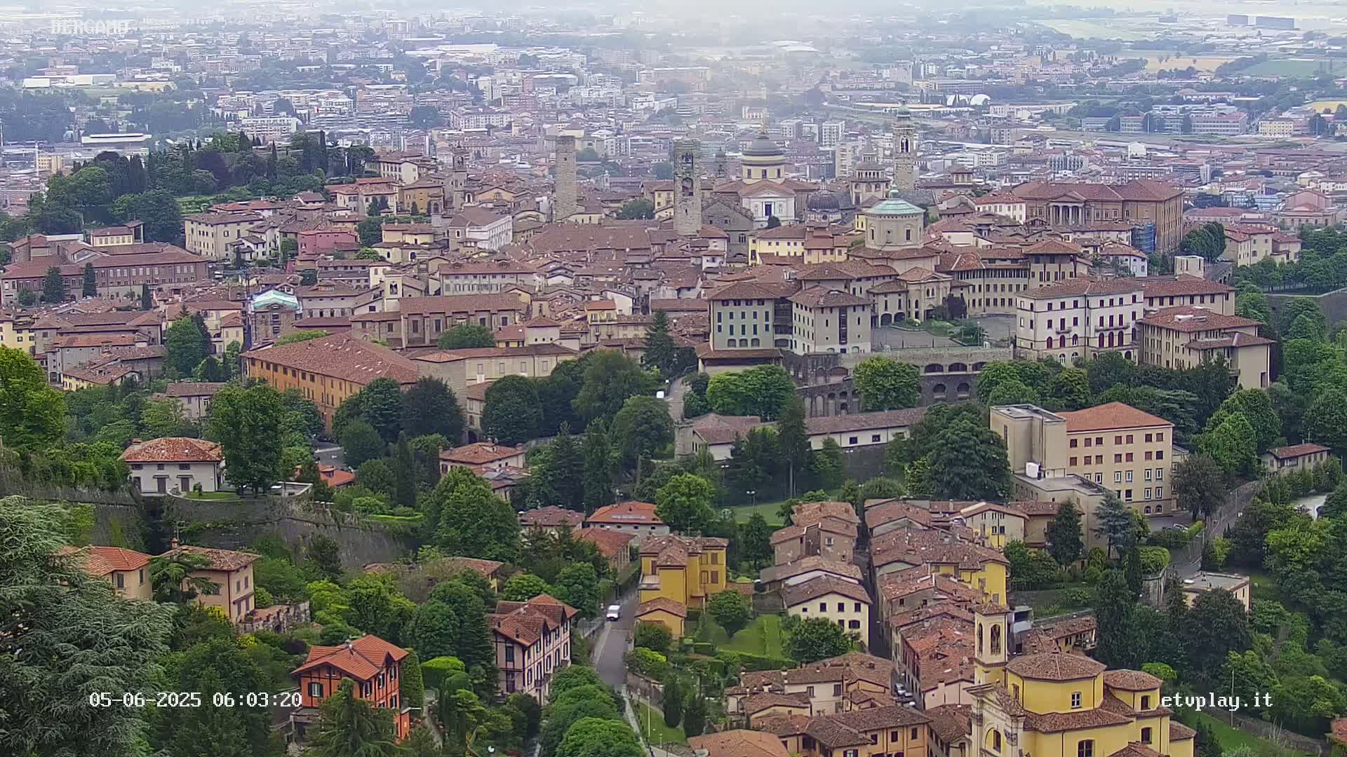 A hazy aerial view shows a densely packed city of low-rise buildings surrounded by lush green trees and a stone wall, nestled within a larger sprawling city.
