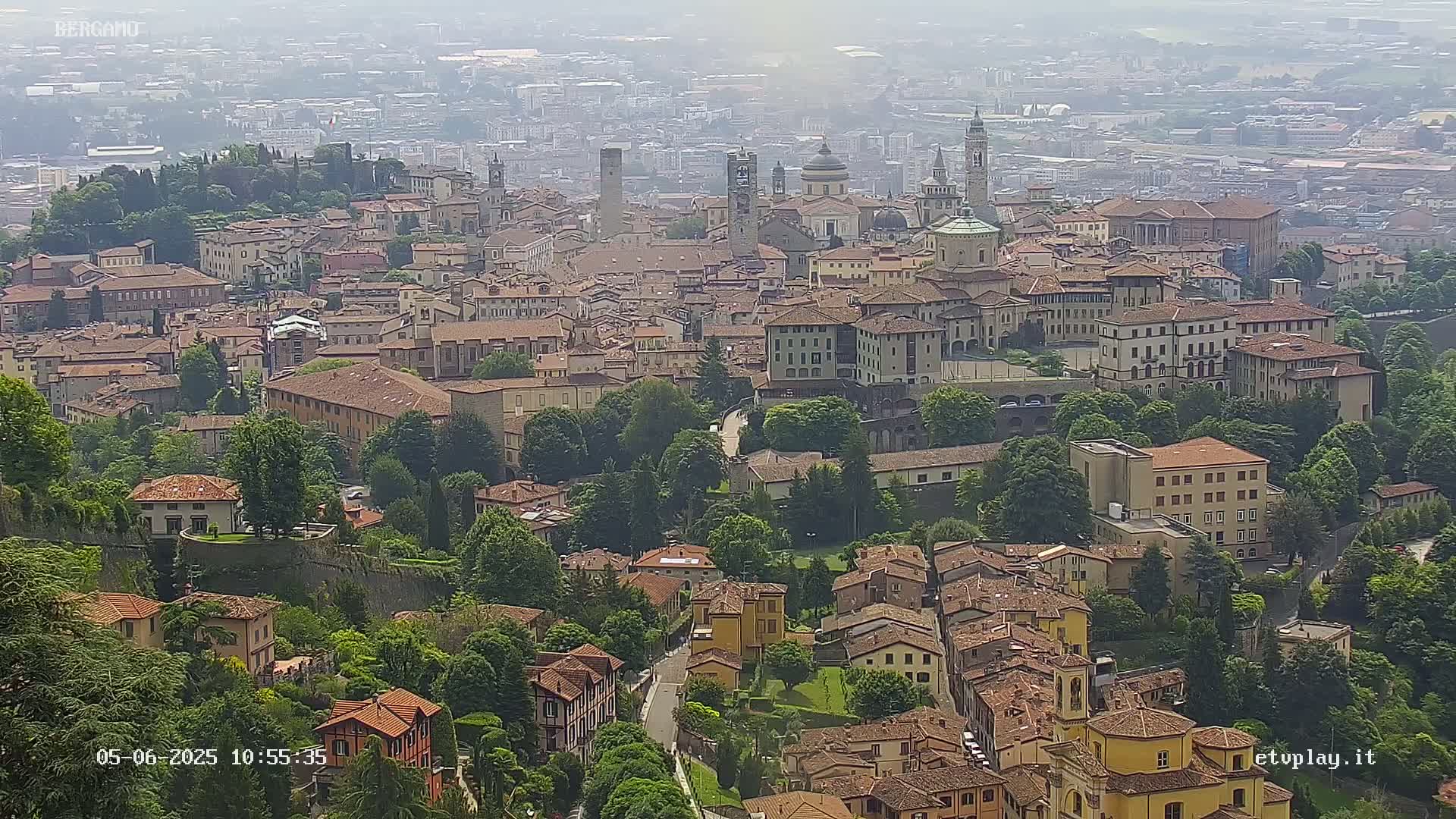 A hazy view from above shows a sprawling city of tan and brown buildings nestled among green trees and hills.