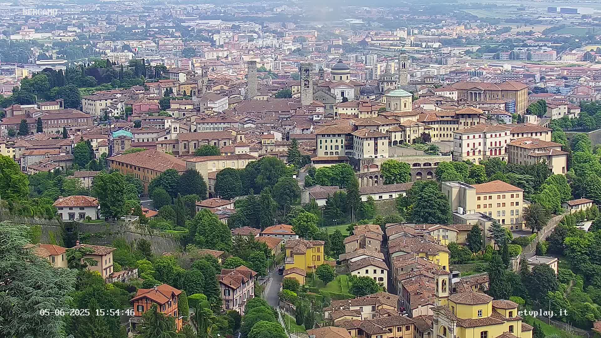 A sprawling city of numerous tan and brown buildings, surrounded by lush green trees and walls, is visible under a hazy sky.