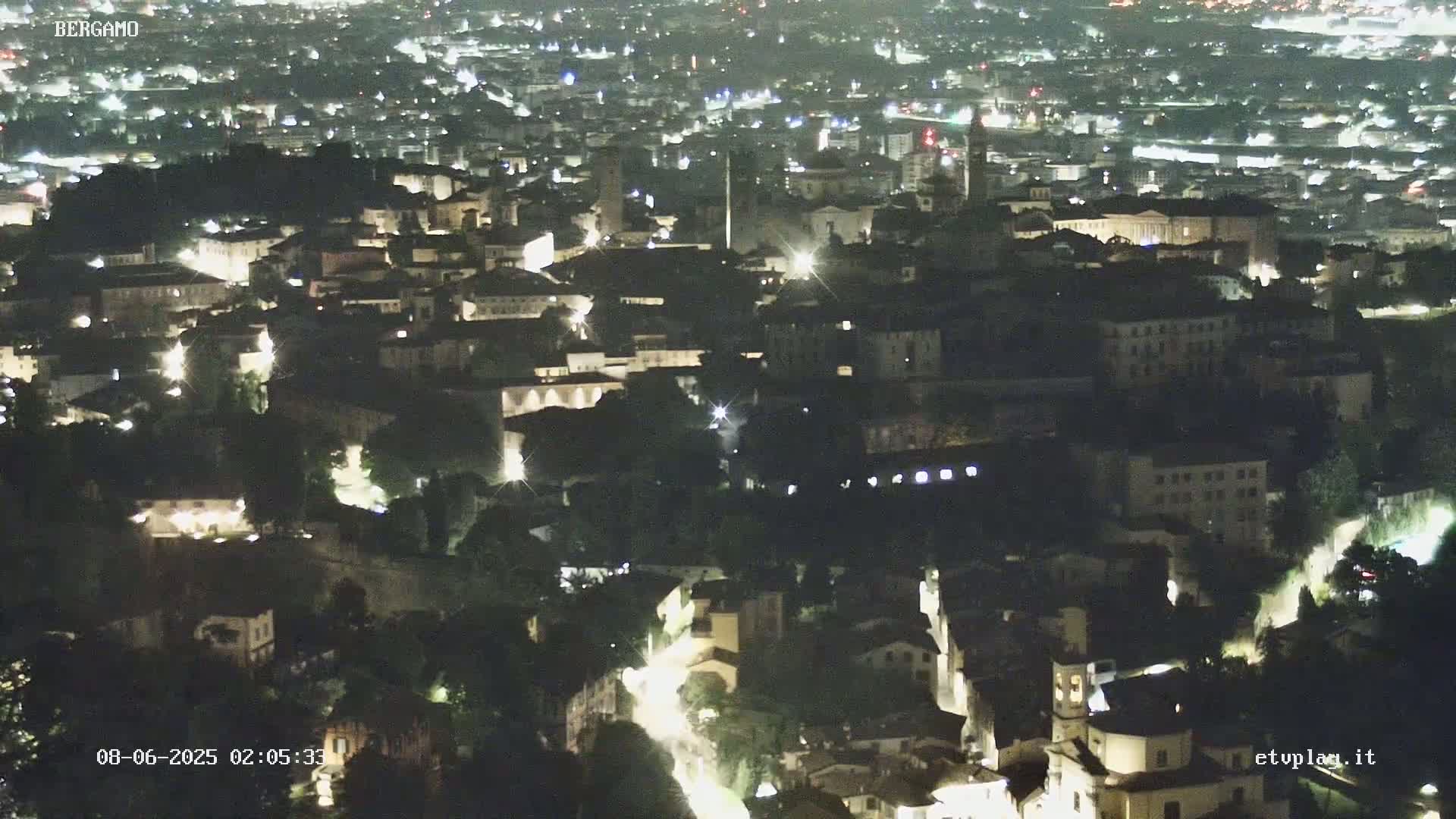 A nighttime aerial view of a city, illuminated by streetlights, showing a dense collection of buildings nestled among greenery.