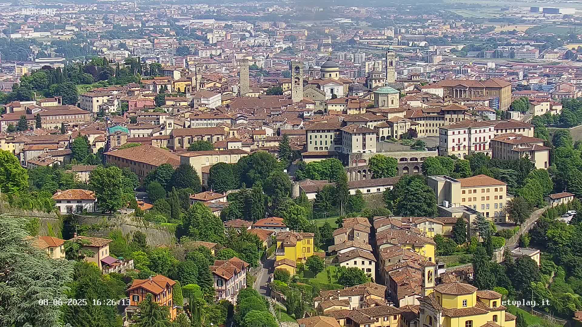 A sprawling city of historic, multi-storied buildings nestled among lush green trees on a sunny day.