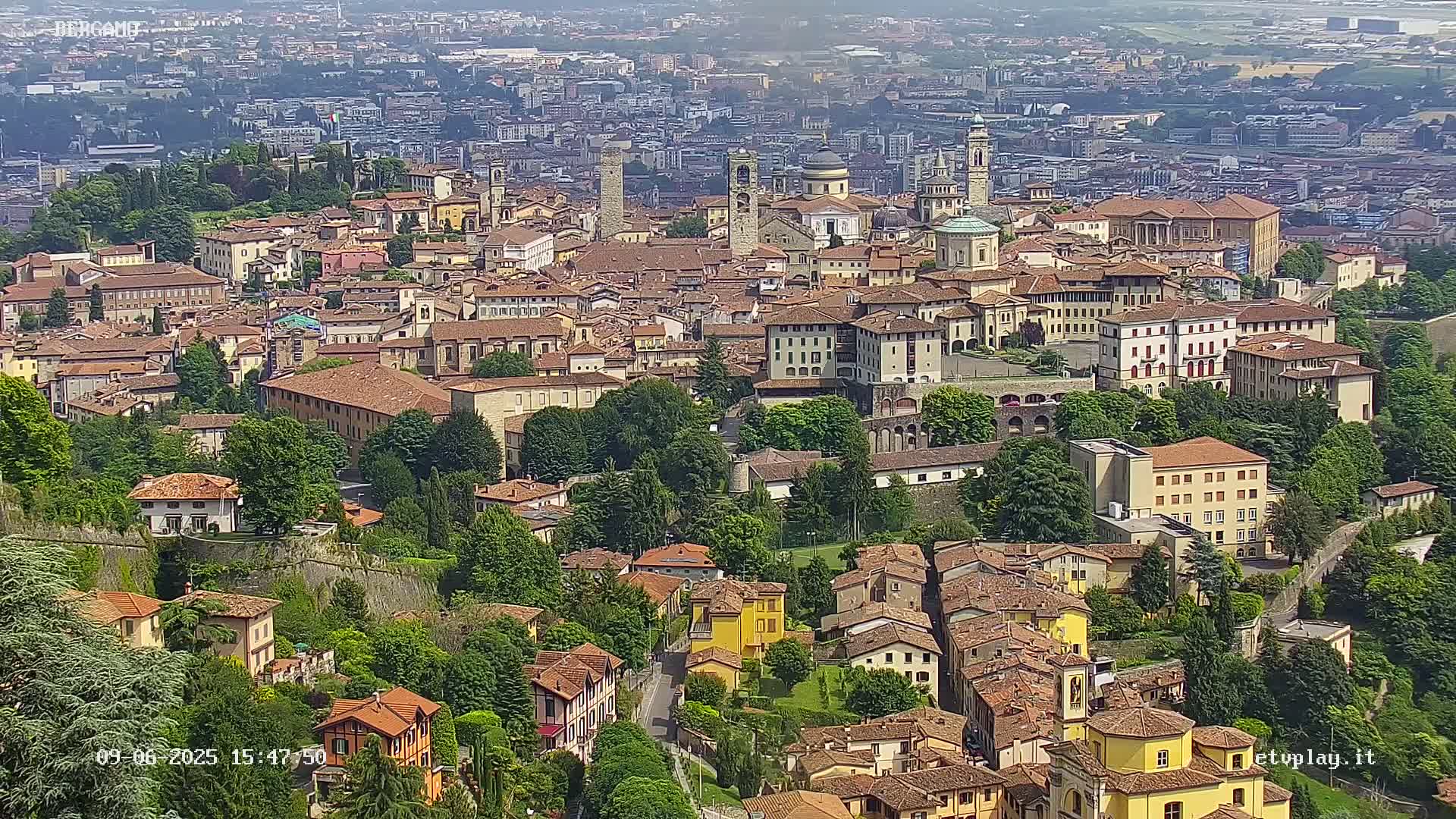A sprawling, historic city with numerous terracotta-roofed buildings is nestled in a verdant valley under a hazy sky.