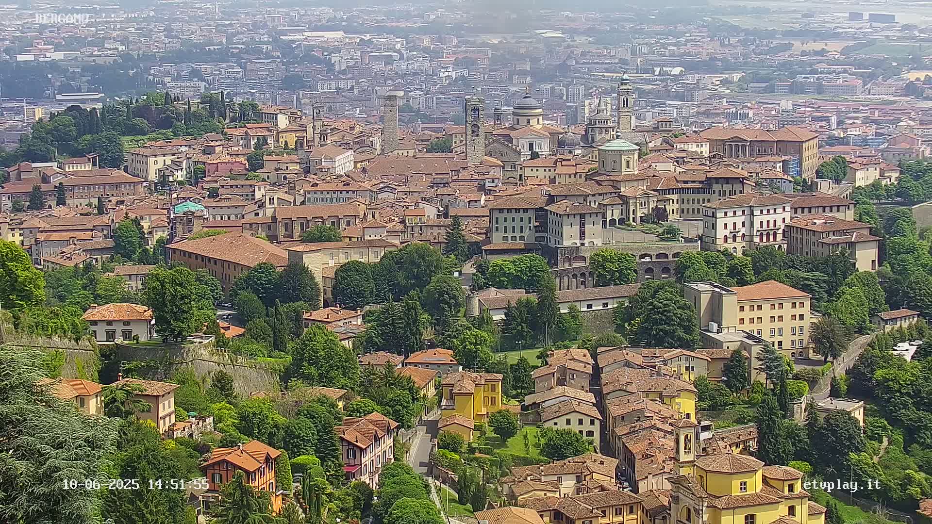 An aerial view shows a large city with many brown-roofed buildings, green trees, and a hazy sky.