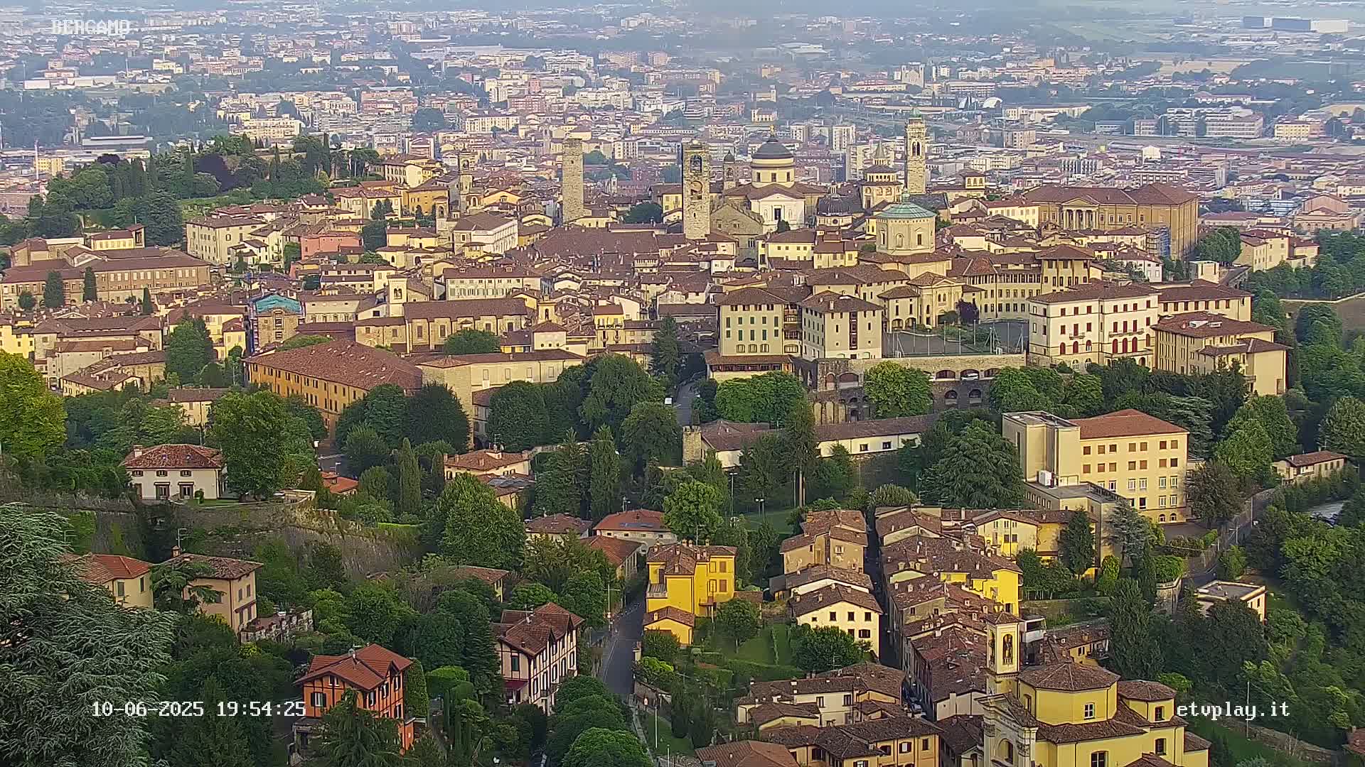A hazy, high-angle view of a densely packed European city, showing many tan and brown buildings nestled amongst lush green trees.