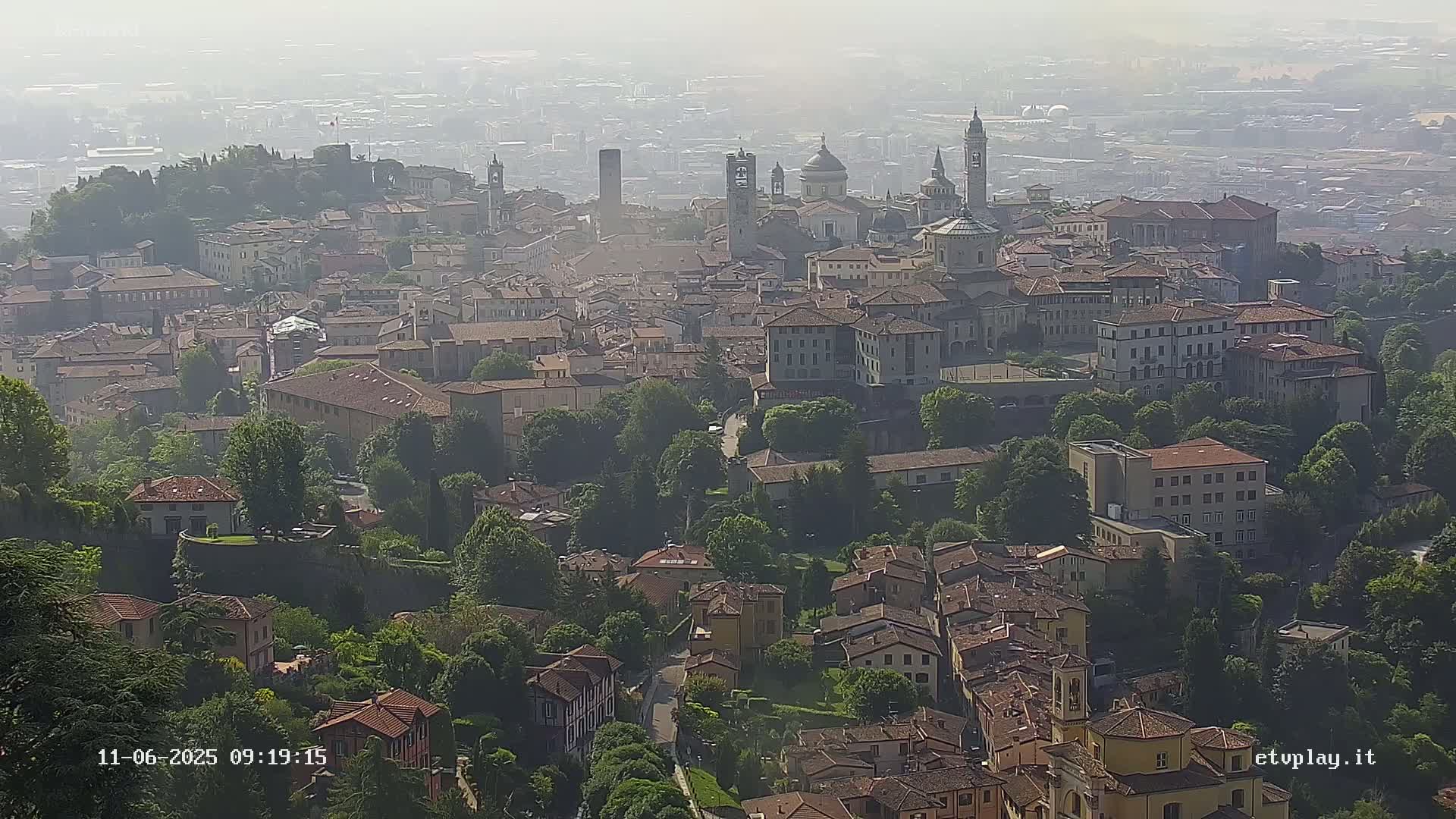 A hazy, high-angle view shows a sprawling city with numerous buildings of varying sizes and architectural styles, nestled among lush green trees.