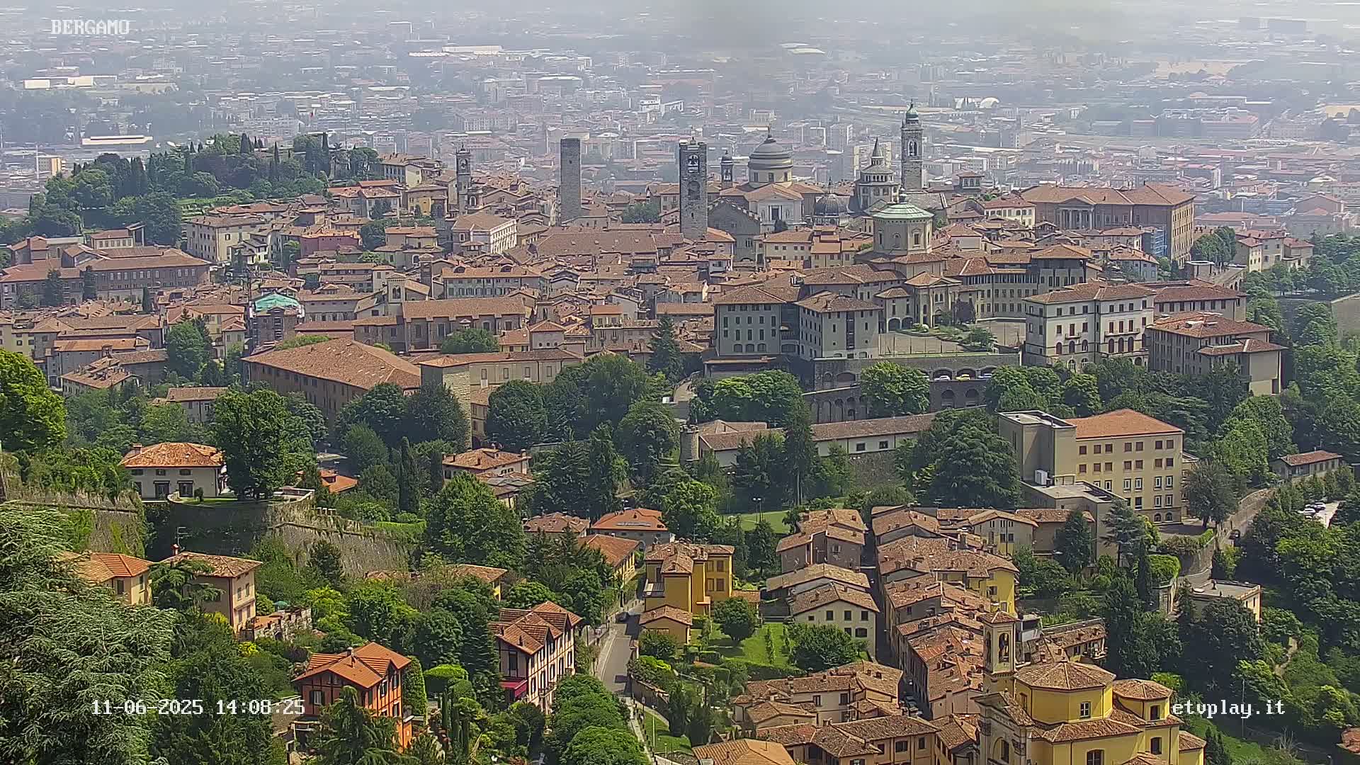 An aerial view shows a hazy, expansive city with numerous terracotta-roofed buildings and green trees under a hazy sky.
