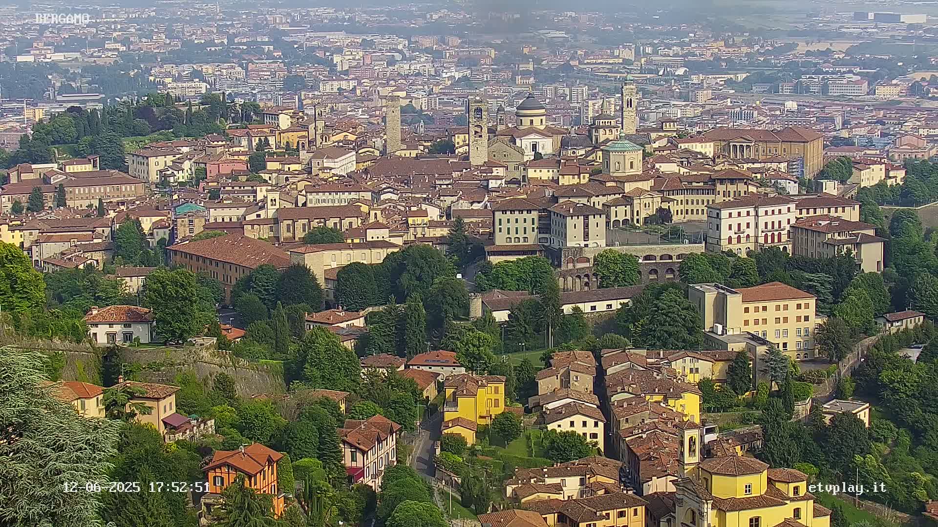 A sprawling city of tan and brown buildings nestled among lush green trees and vegetation under a hazy sky.