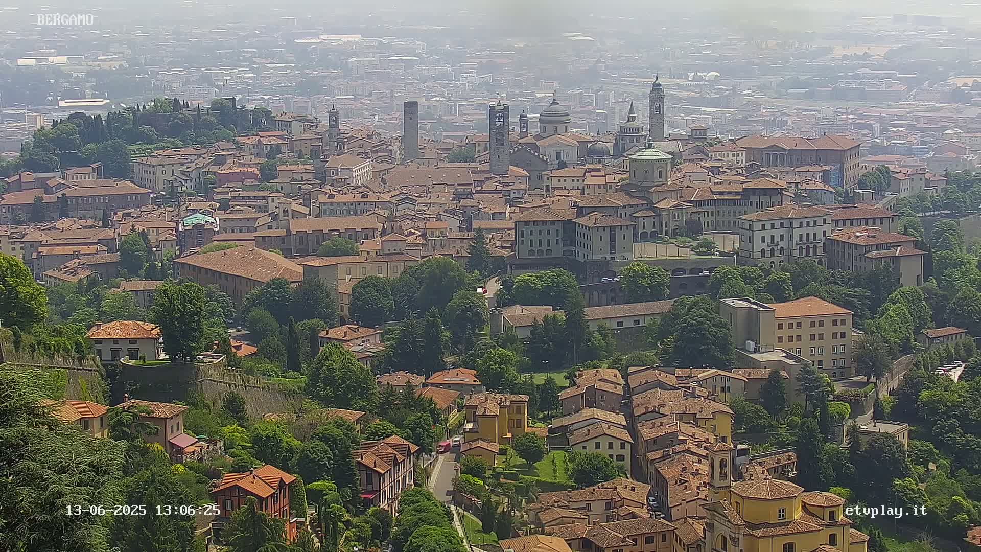 A hazy view of a city with numerous red-roofed buildings, several towers, and green spaces, all nestled on a hillside.