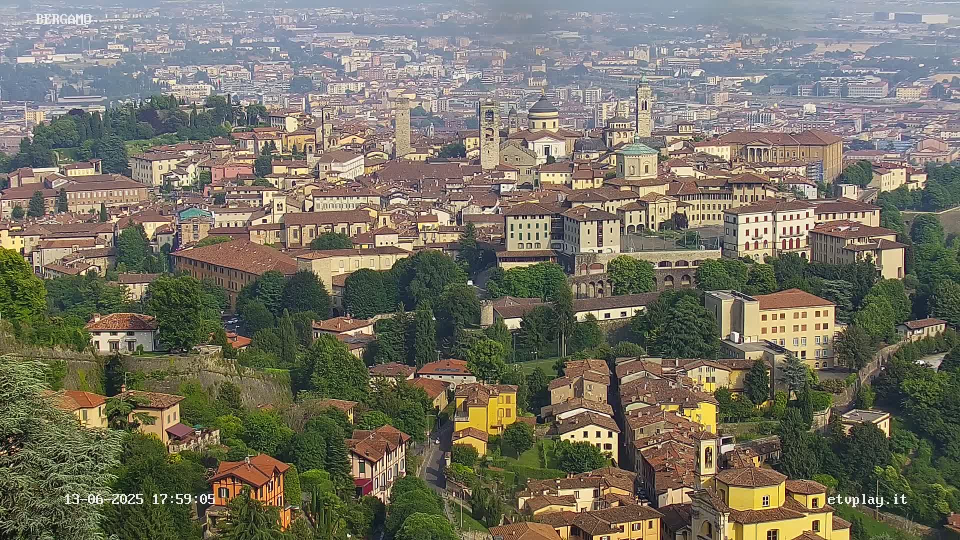 A hazy, high-angle view shows a sprawling city of tan and red-roofed buildings nestled among green hills.