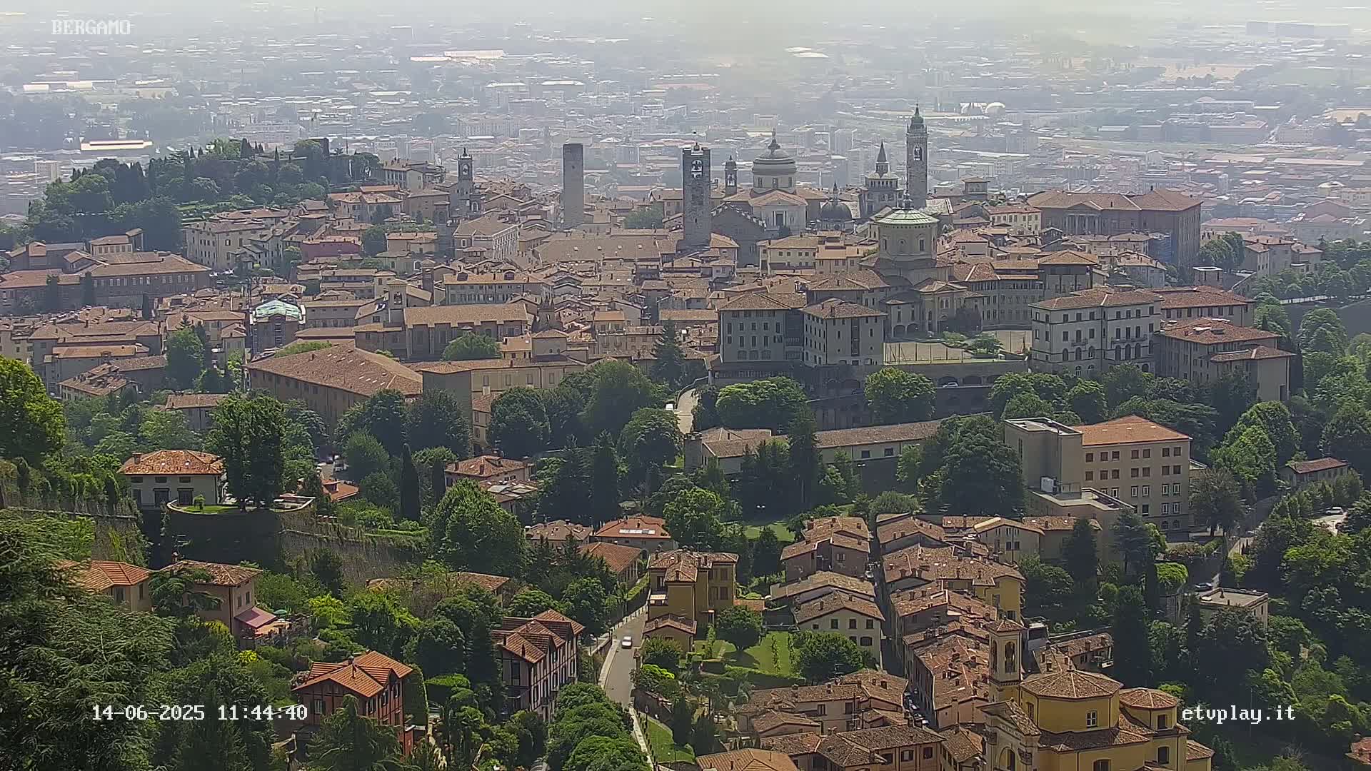 A hazy, high-angle view shows a large city with many brown-roofed buildings, interspersed with green trees and surrounded by a wall.