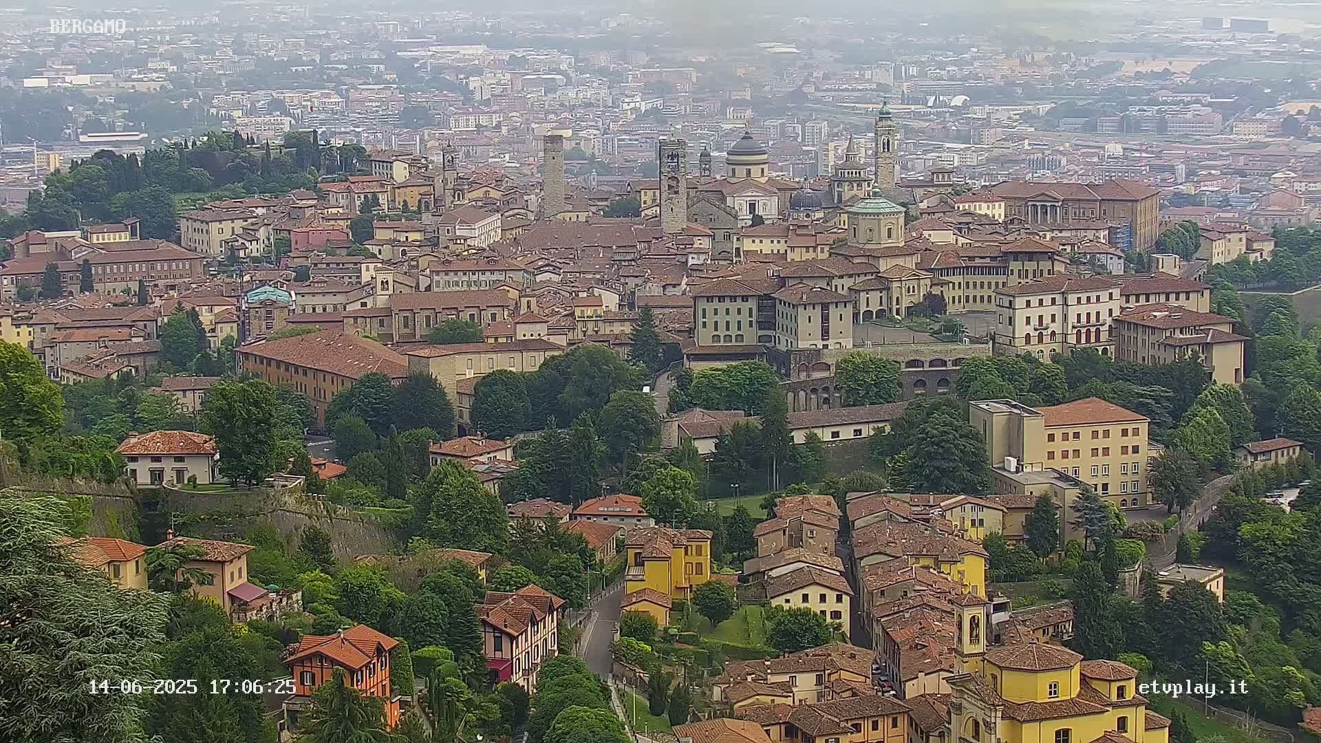 A hazy view from above shows a sprawling city of closely packed buildings with red-tiled roofs, interspersed with green trees and walls, under a cloudy sky.
