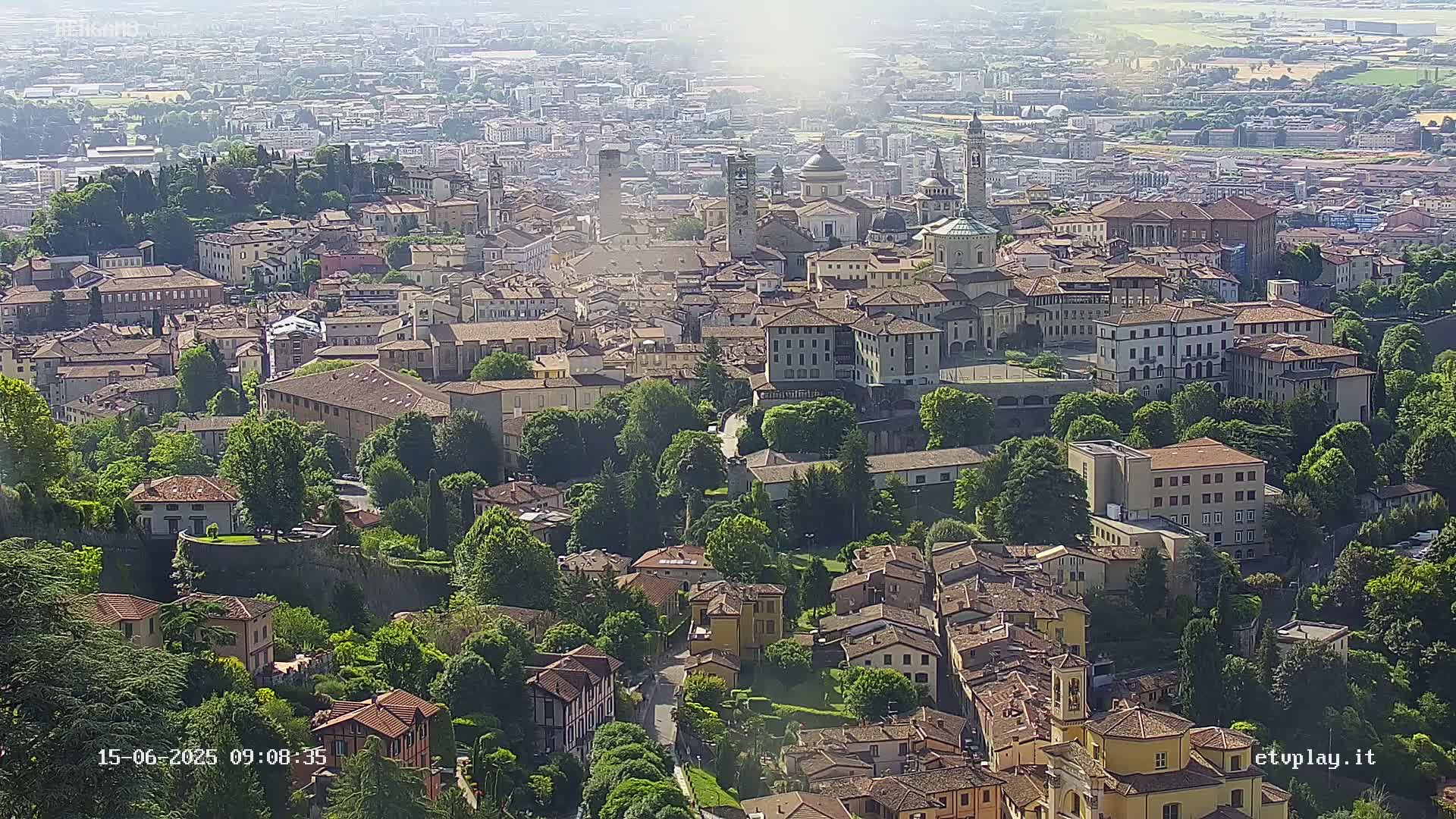 A sprawling city with numerous buildings nestled amongst lush green trees under a hazy, sunny sky.