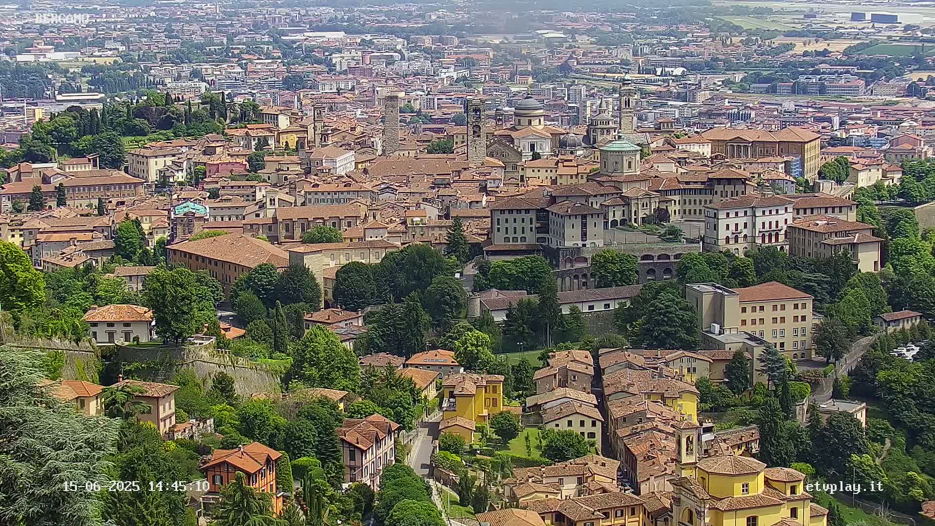 A sprawling city of terracotta-roofed buildings nestled among lush green trees and hills under a clear, sunny sky.