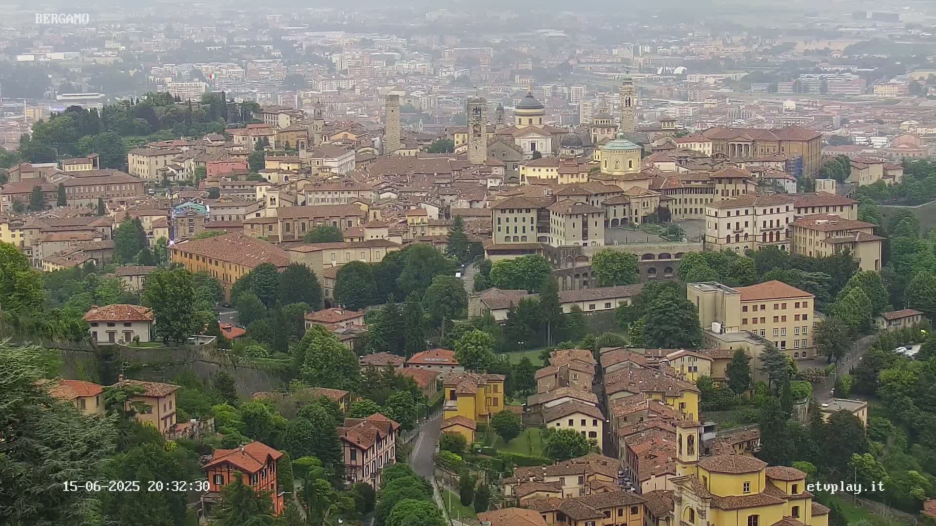 A hazy, high-angle view shows a dense collection of buildings in a European city, nestled amongst lush green trees and foliage.