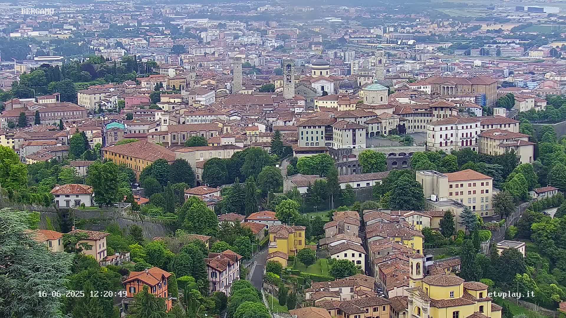 A sprawling town of closely-packed, low-rise buildings with red-tiled roofs, interspersed with green trees and surrounded by a stone wall, is visible under an overcast sky.
