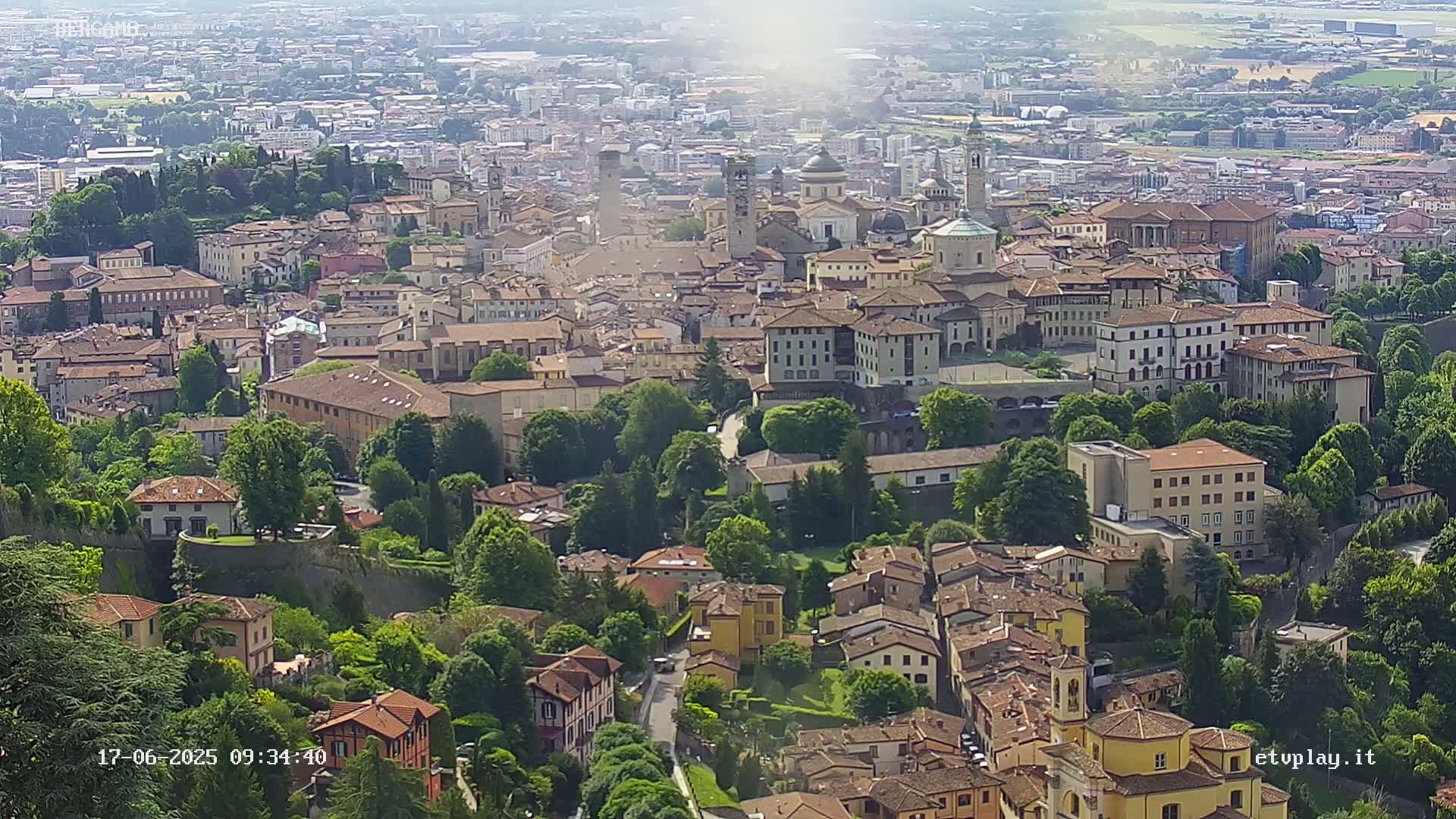 An aerial view of a city with numerous buildings, surrounded by lush green trees and foliage under a bright, hazy sky.