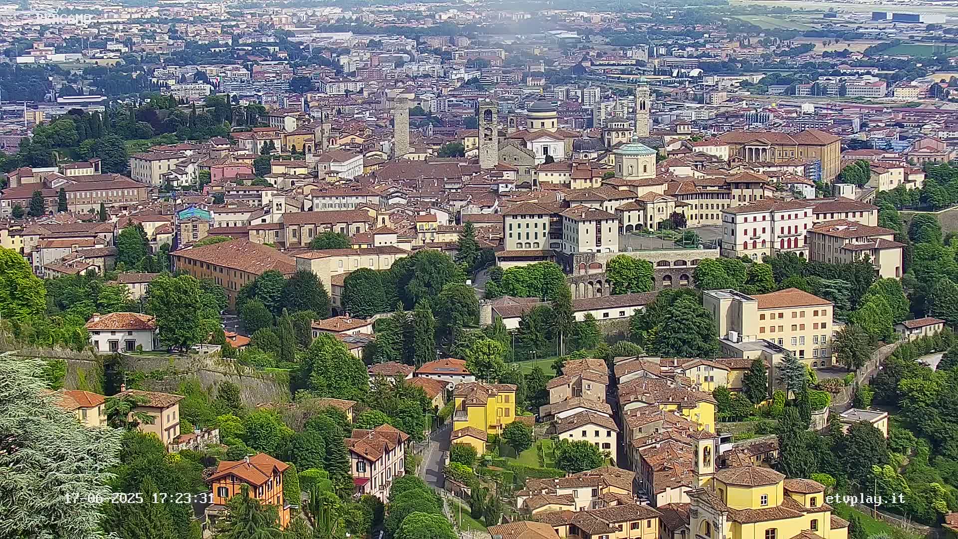 A sprawling city of tan and brown buildings with red-tiled roofs is nestled among green trees under a mostly clear sky.