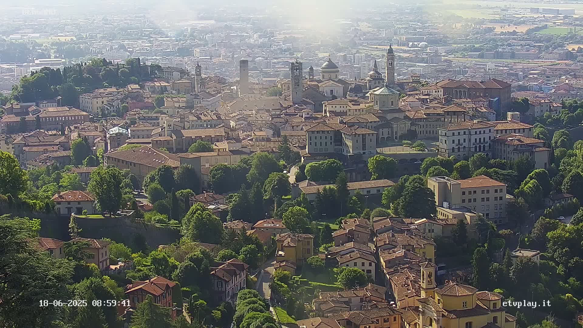 A hazy, sunny day reveals an aerial view of a sprawling town nestled among lush green hills, with numerous buildings of varying sizes and architectural styles.