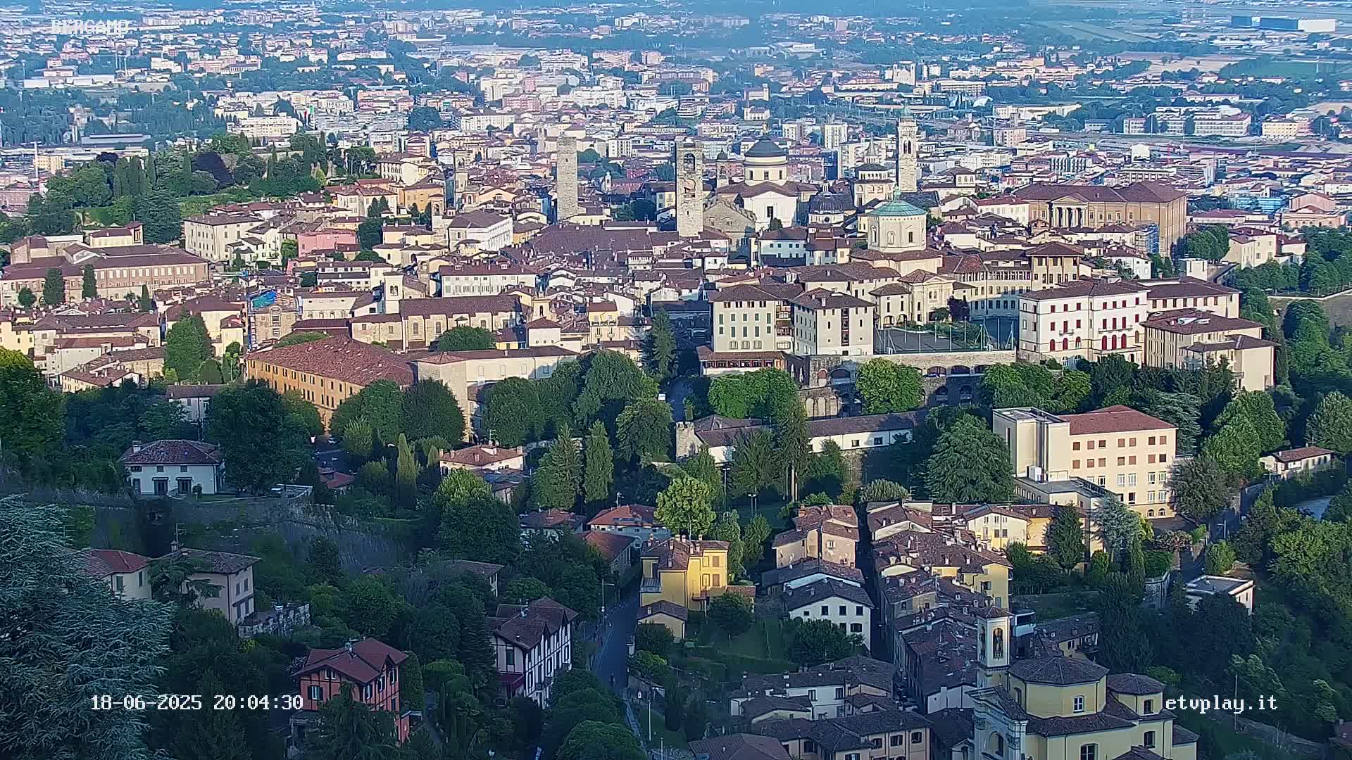 An aerial view shows a sprawling city with numerous buildings of varying sizes and architectural styles, nestled among lush green trees under a hazy sky.