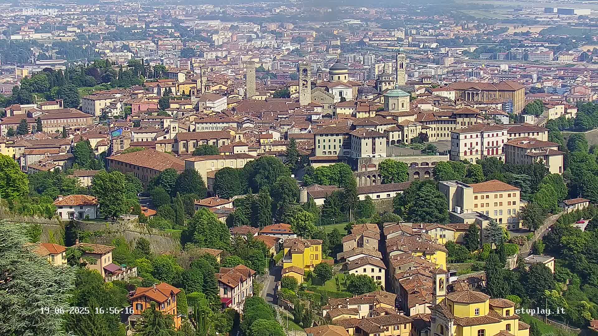 An aerial view of a sprawling city on a sunny day, showing a dense collection of historic buildings surrounded by lush green trees and walls.