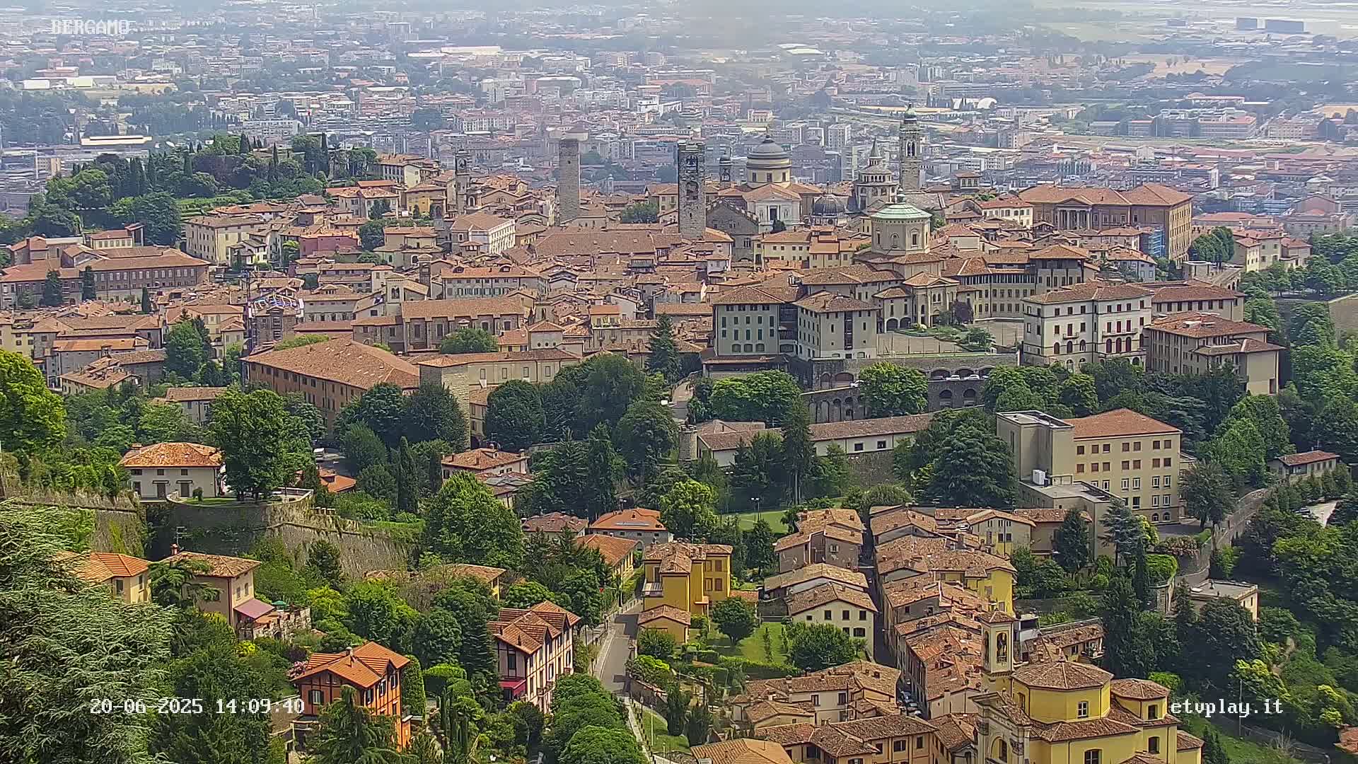 A hazy, high-angle view of a dense collection of red-roofed buildings nestled in a lush green valley, with a sprawling city visible in the background.