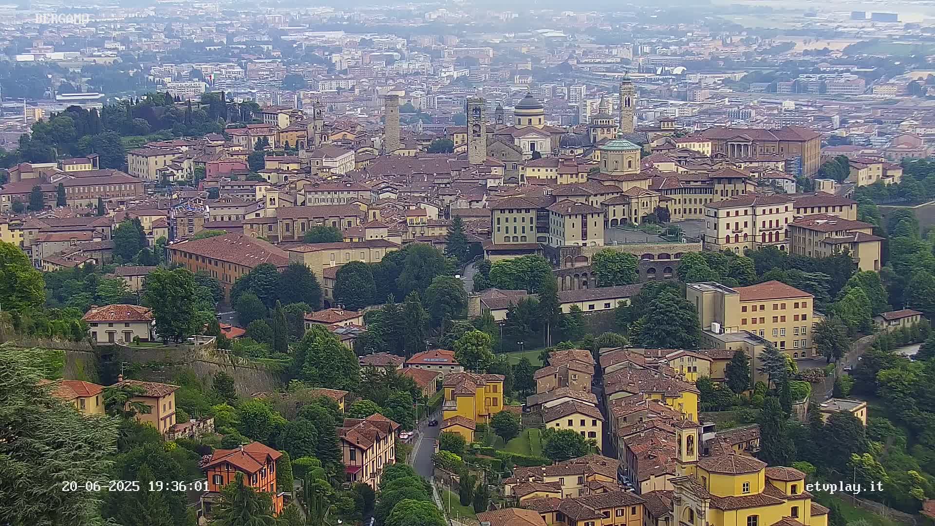 A hazy, high-angle view of a dense collection of historic buildings in a European city, nestled amongst green trees and surrounded by more distant buildings.