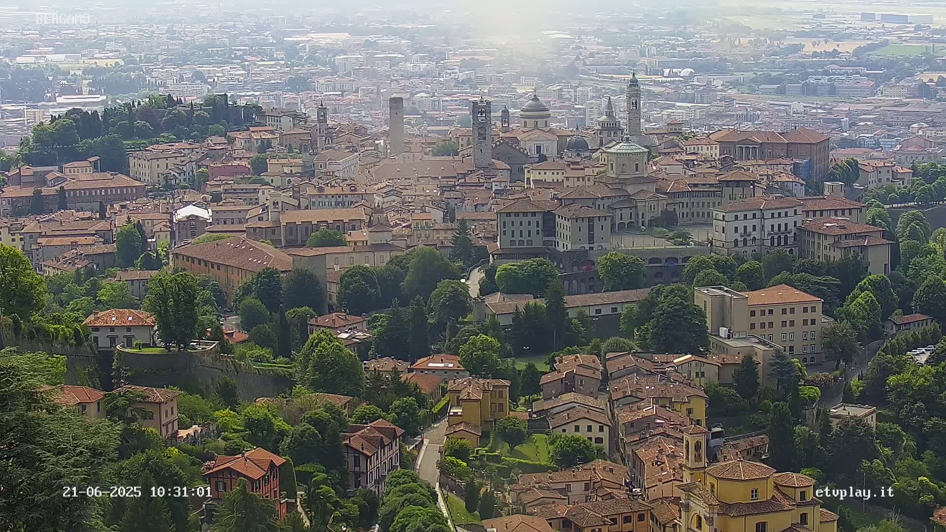 A hazy, sunny day reveals a sprawling cityscape of low-rise buildings and churches nestled among lush green trees and hills.