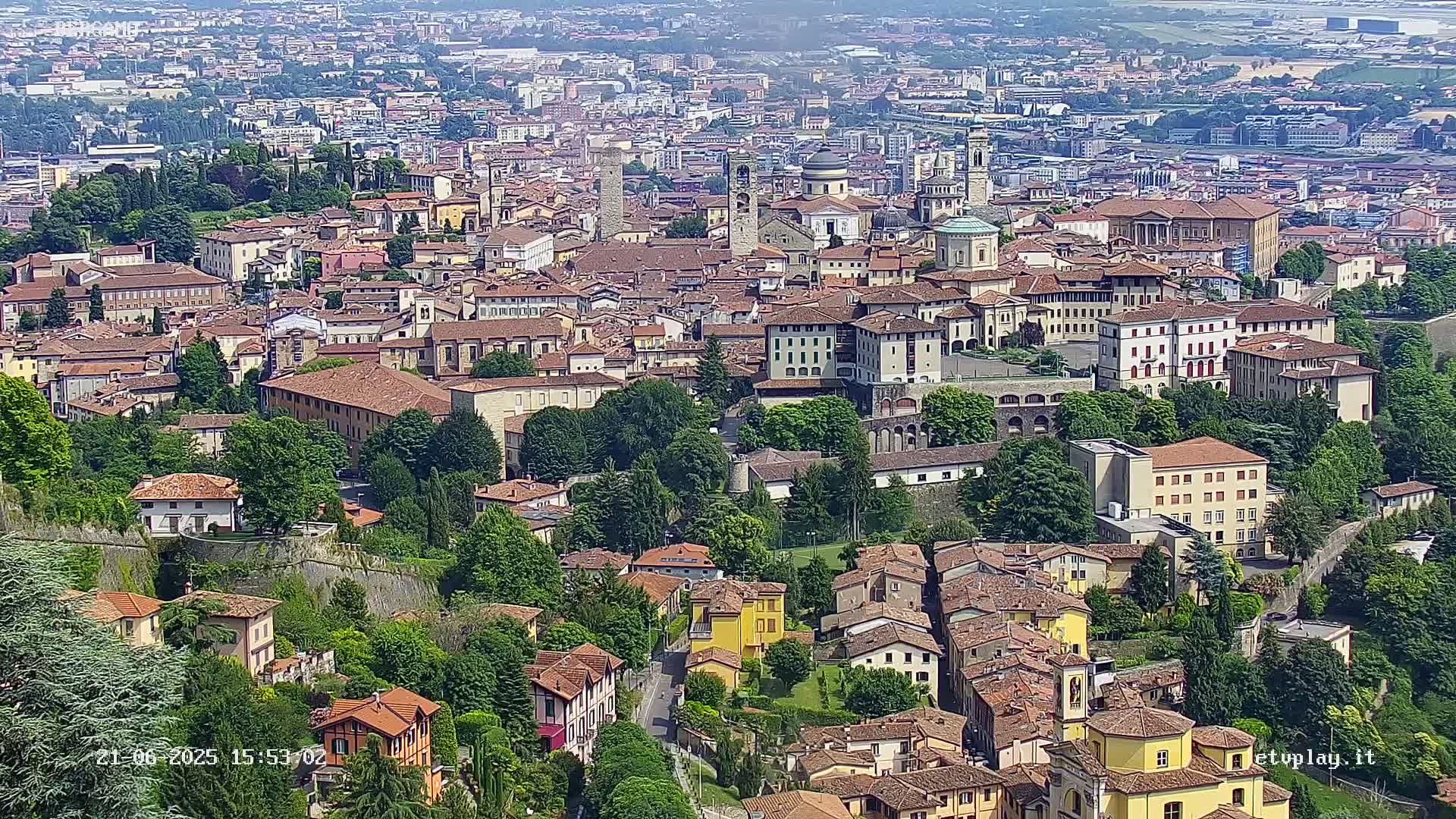 A sprawling city of terracotta-roofed buildings, nestled amongst lush green trees and walled sections, is visible under a clear sky.