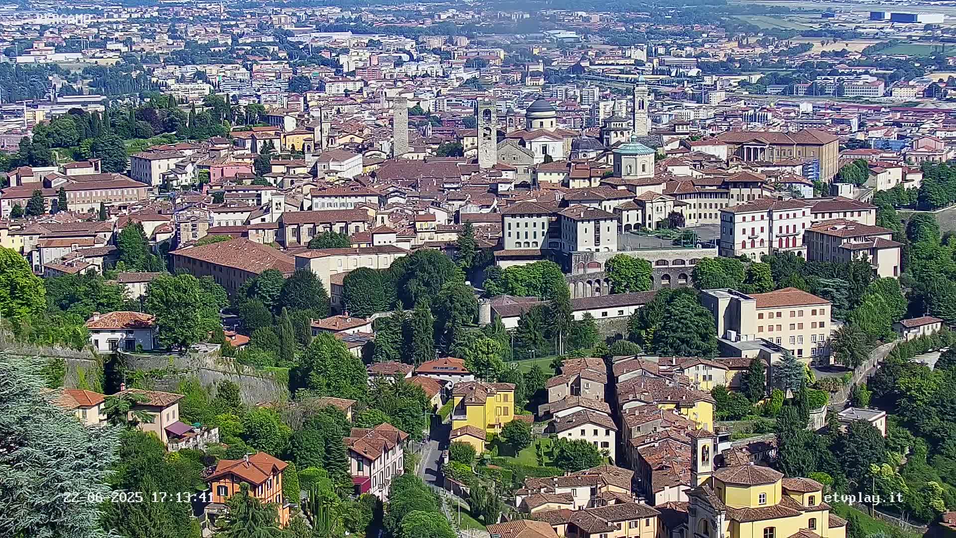 A sprawling city with many brown-roofed buildings, surrounded by lush green trees, under a bright, sunny sky.