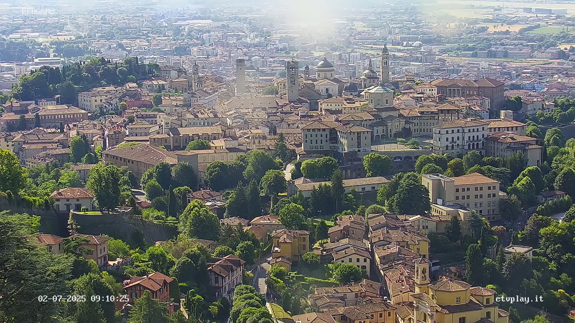 A sprawling city with numerous buildings of varying heights and architectural styles, nestled amongst lush green trees under a bright, sunny sky.