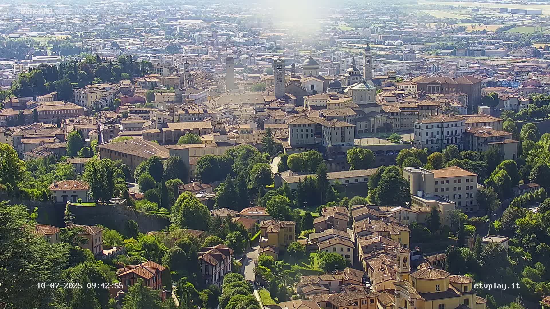 A sprawling city with many brown-roofed buildings, nestled amongst lush green trees, under a bright, sunny sky.