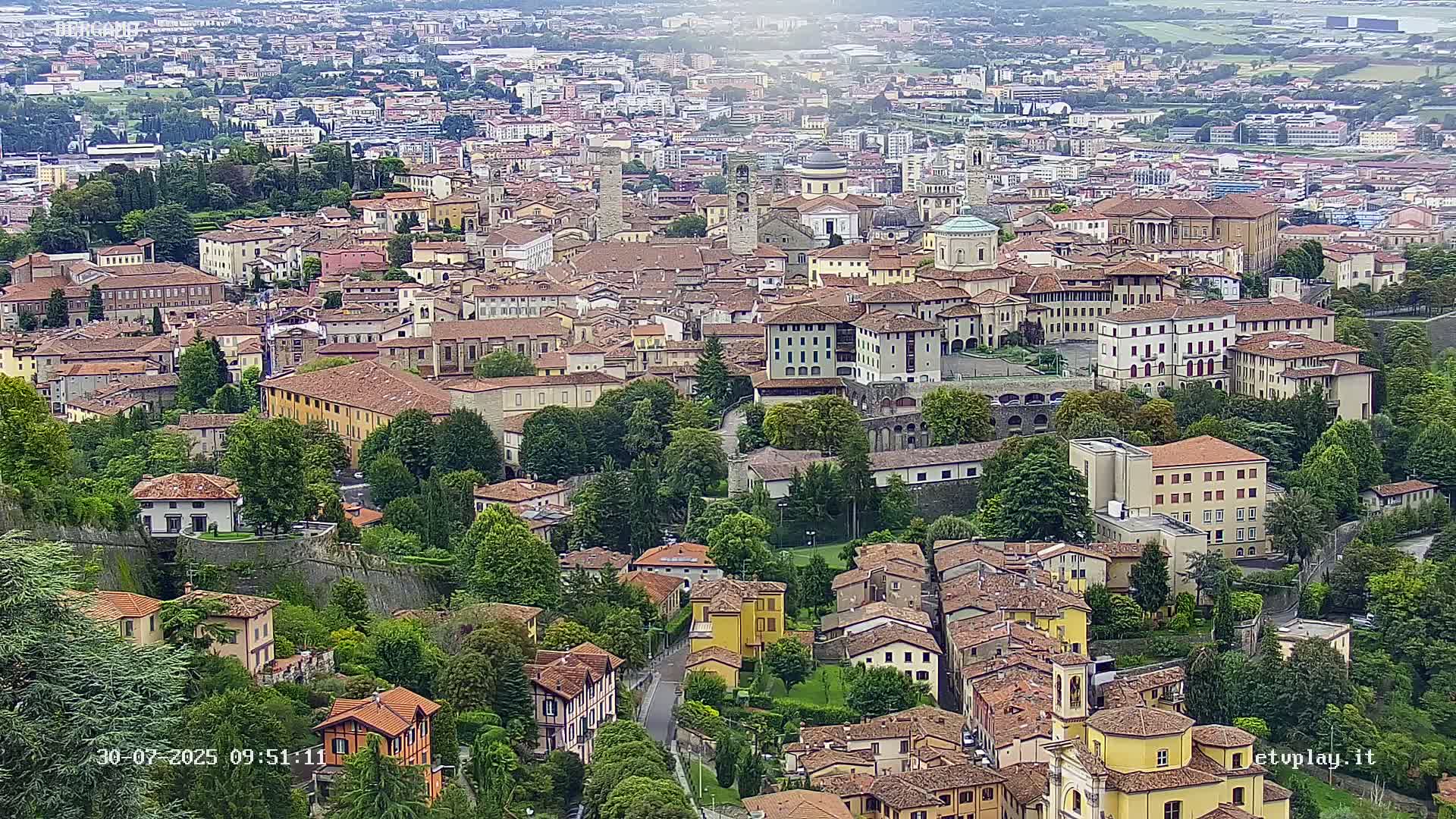 A sprawling city of low-rise, terracotta-roofed buildings sits nestled among lush green trees under a mostly cloudy sky.
