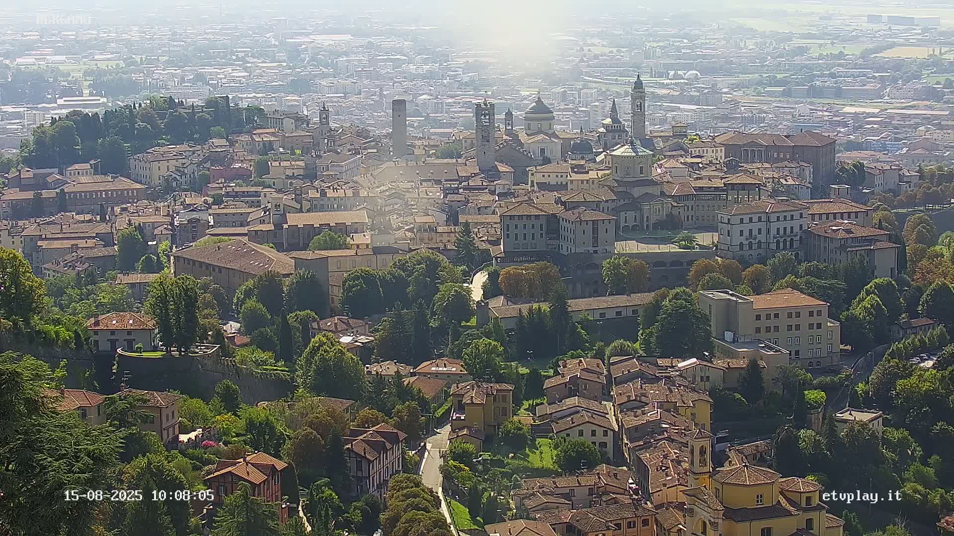 A hazy, sunny day reveals an aerial view of a sprawling European town, characterized by densely packed buildings, churches with steeples, and green spaces nestled amongst the architecture.