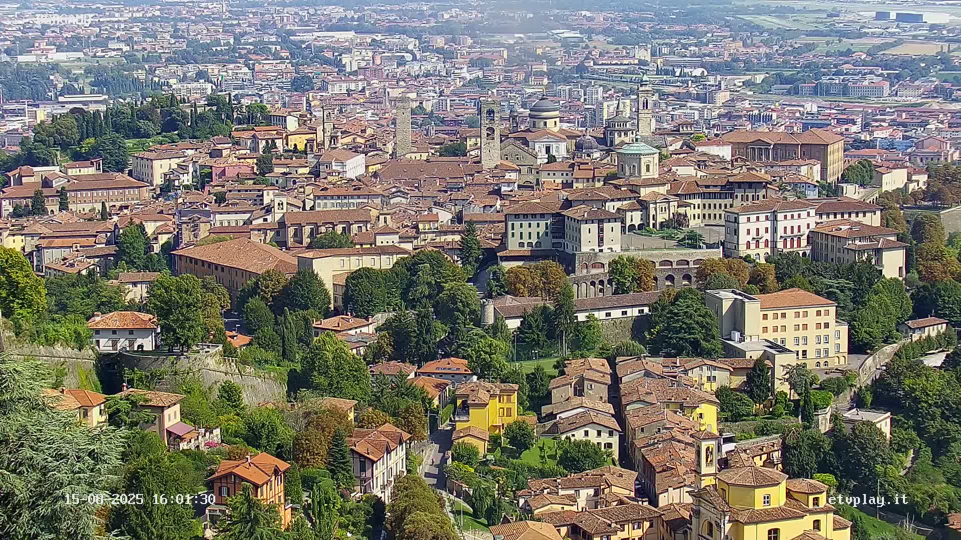 A hazy, sunny day reveals an aerial view of a sprawling European town, characterized by densely packed buildings, churches with steeples, and green spaces nestled amongst the architecture.