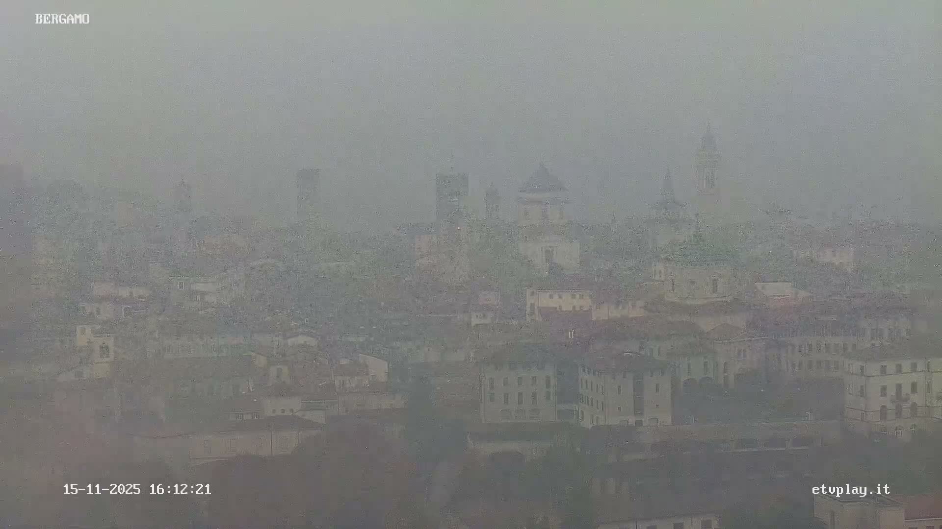A densely built cityscape with prominent domes and bell towers is heavily obscured by thick fog, creating extremely low visibility and a muted, gray atmosphere.