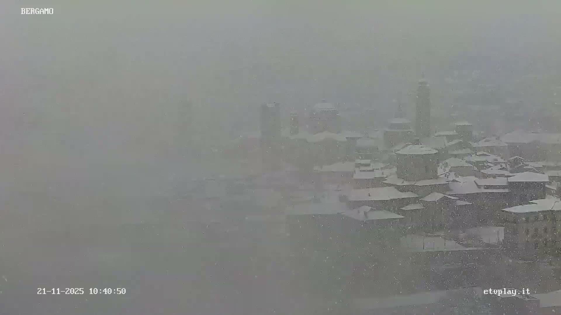 An aerial view reveals a snow-covered town with buildings and towers partially obscured by heavy, falling snow.