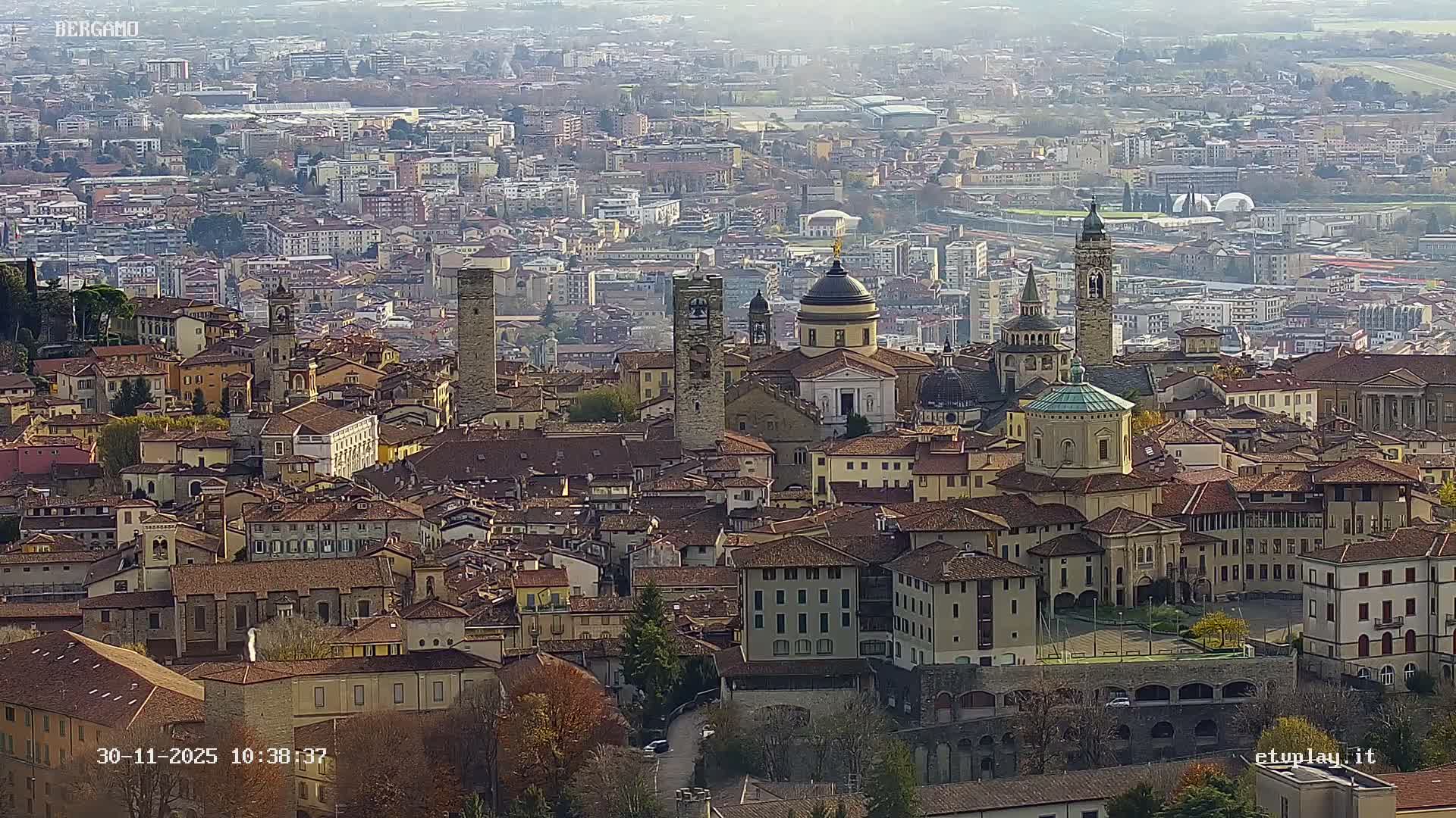 A sprawling European city, characterized by a mix of historic buildings with domes and bell towers amidst dense housing and autumn trees, is seen under a hazy, overcast sky.