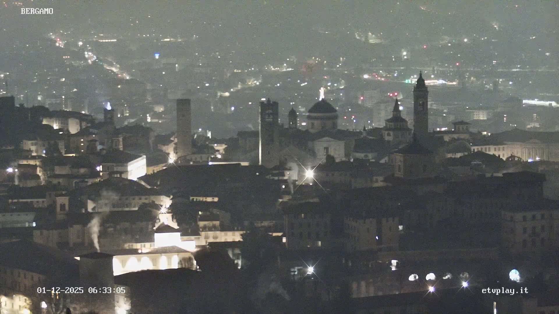 A hazy night view reveals a sprawling, illuminated city featuring numerous buildings, prominent church domes, and bell towers, all fading into the misty distance.