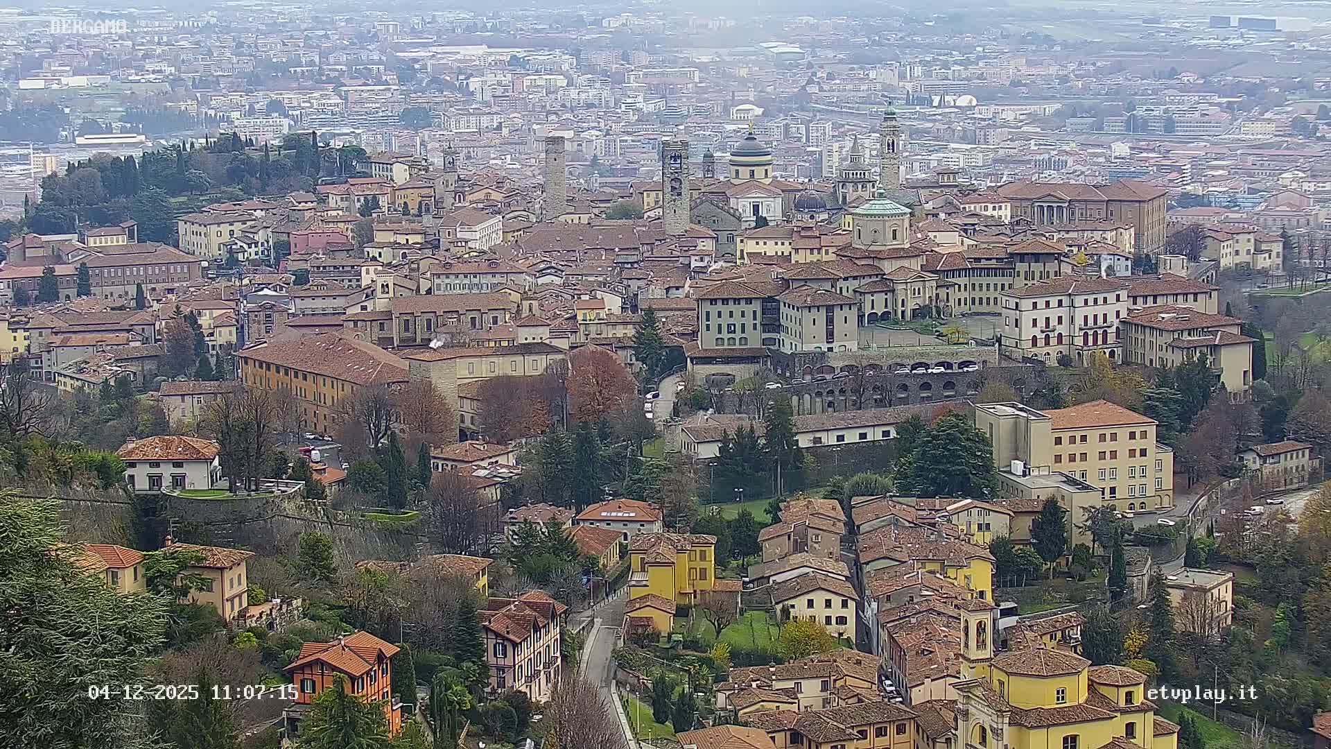 An expansive elevated view of a European city features terracotta-roofed buildings, domes, and bell towers clustered across hilly terrain interspersed with evergreen and bare deciduous trees, all under a hazy, overcast sky.
