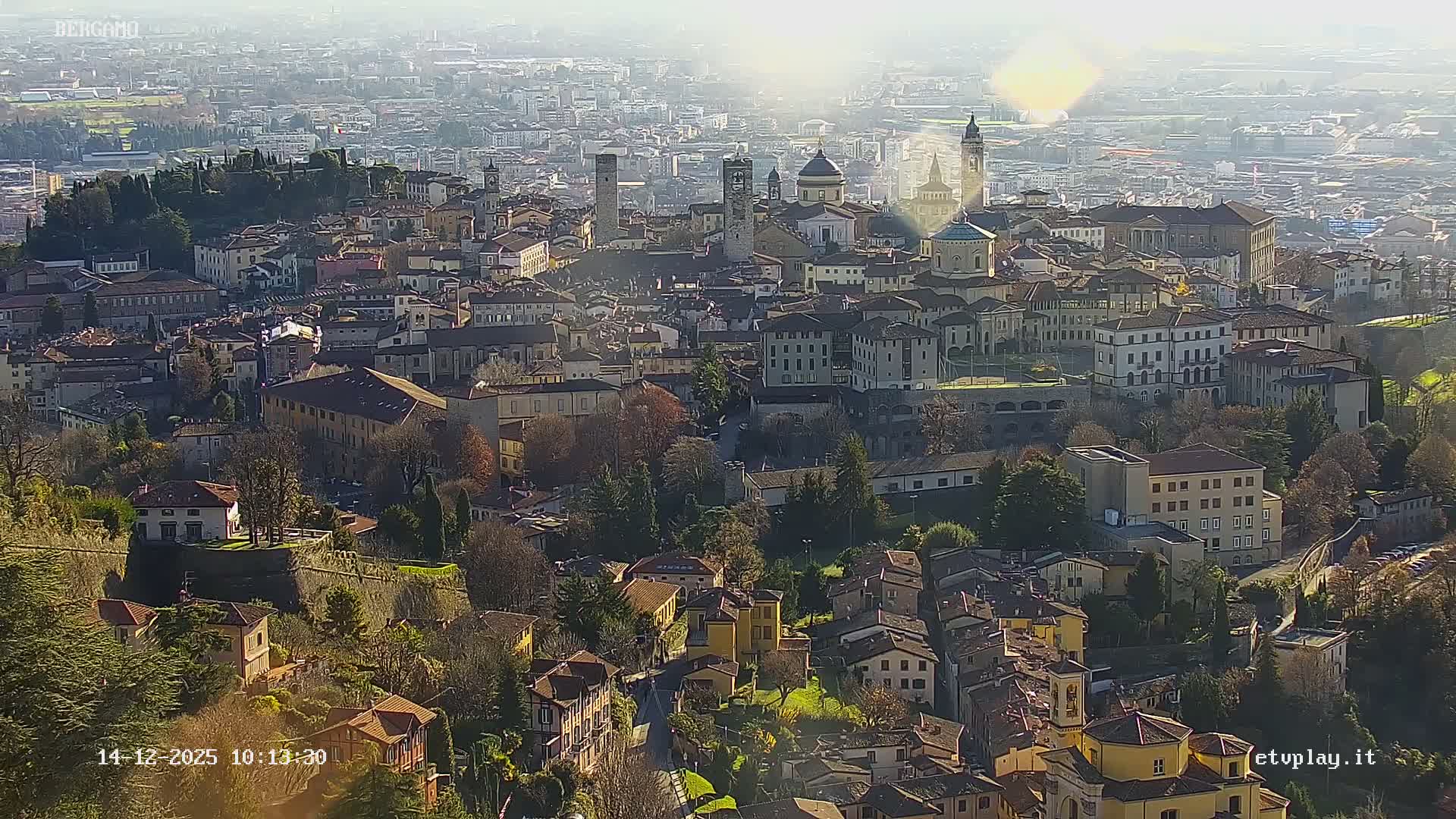 An expansive elevated view of a European city features terracotta-roofed buildings, domes, and bell towers clustered across hilly terrain interspersed with evergreen and bare deciduous trees, all under a hazy, overcast sky.