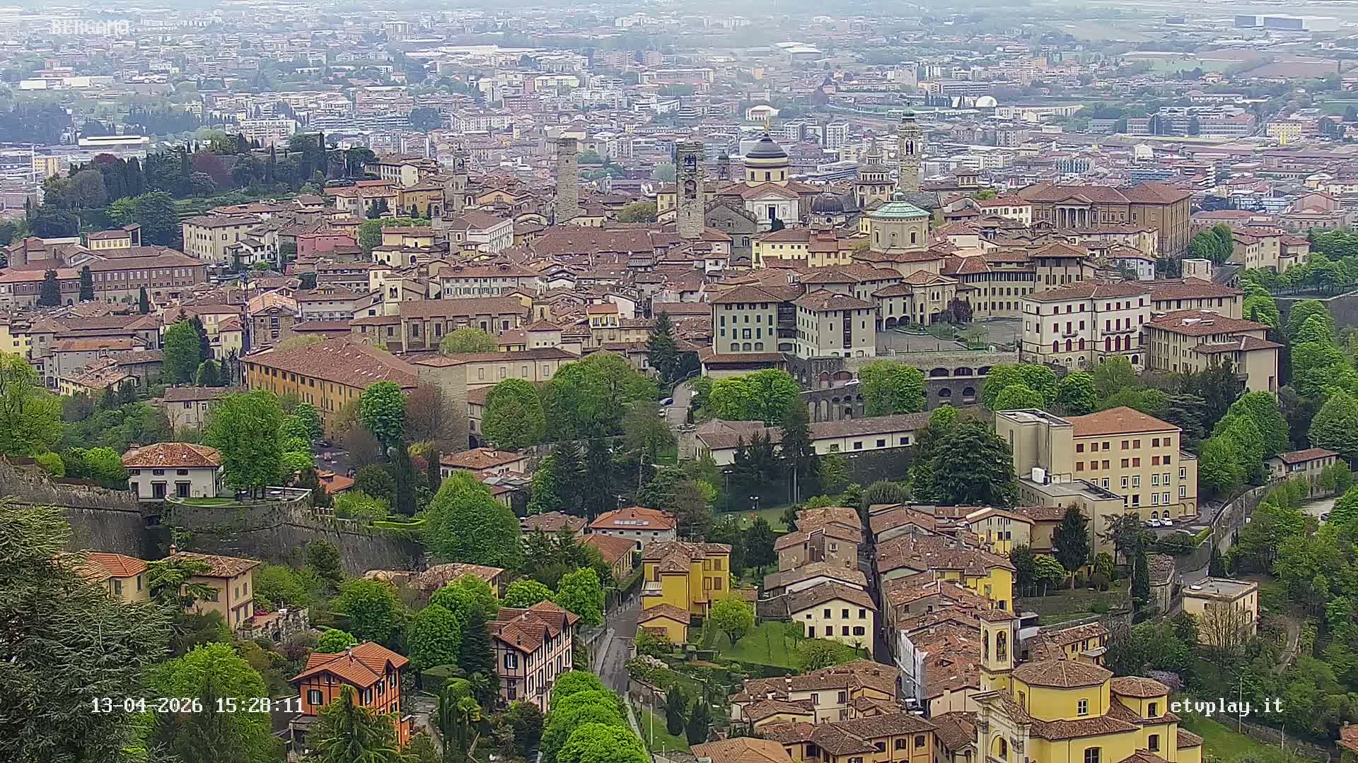 An expansive elevated view of a European city features terracotta-roofed buildings, domes, and bell towers clustered across hilly terrain interspersed with evergreen and bare deciduous trees, all under a hazy, overcast sky.