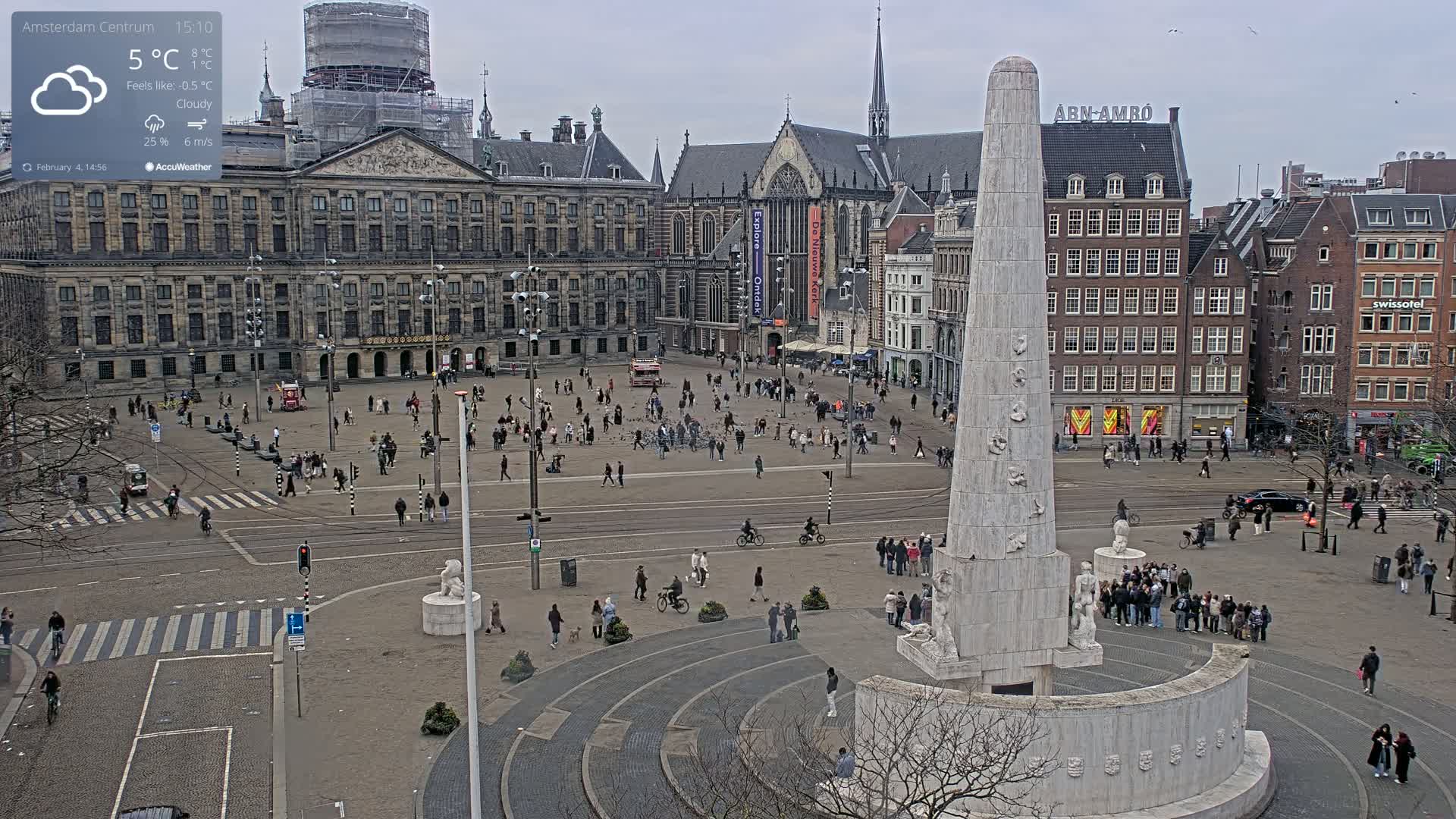 A cloudy day in a large European city square, featuring a tall monument in the foreground, surrounded by historical buildings and people walking around.