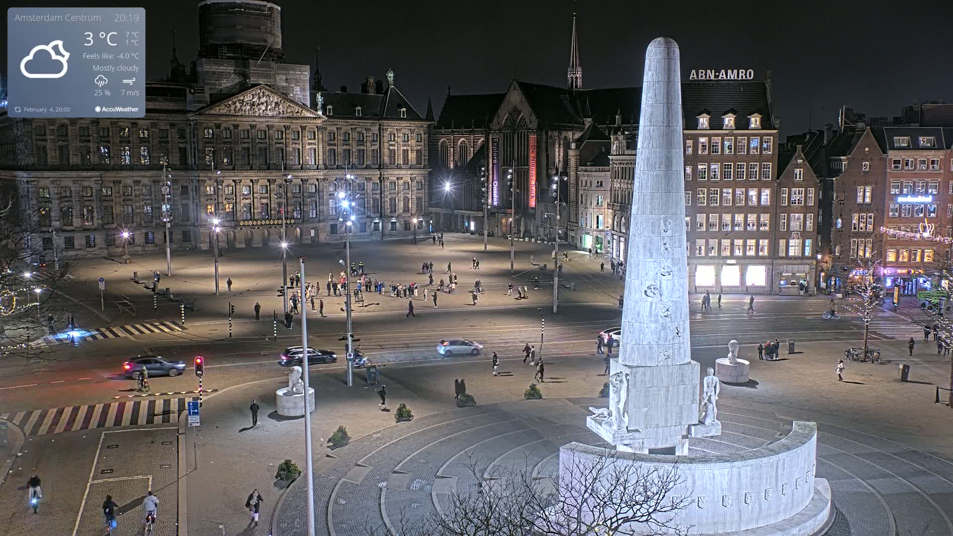 A nighttime, mostly cloudy view of a city square with a tall monument at its center, surrounded by buildings and people.