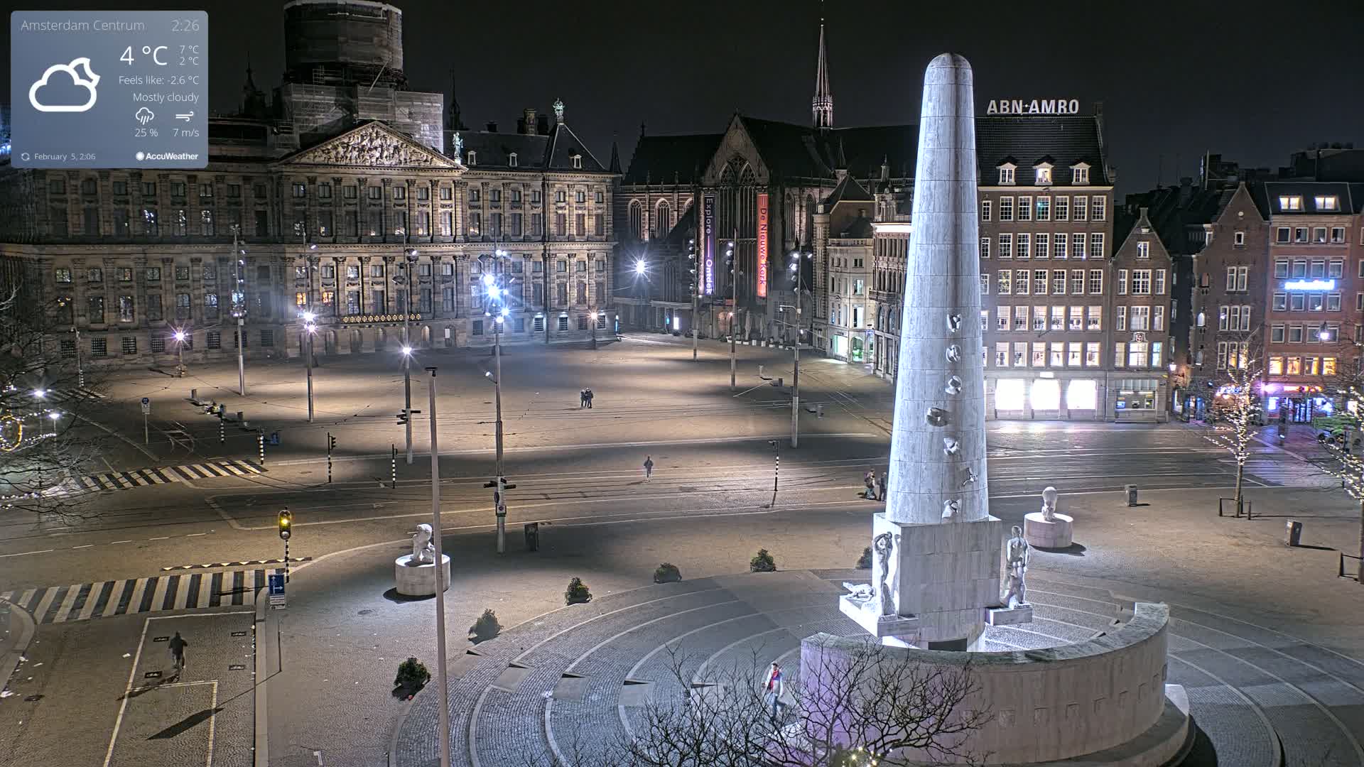 A mostly cloudy night scene shows a large, empty town square with a tall monument in the foreground and several buildings surrounding the square, some of which are lit.